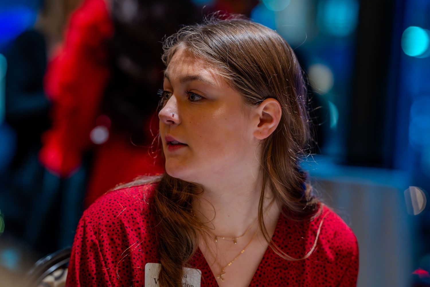 Young woman with braided brown hair wearing a red sweater and gold necklace in a softly lit indoor setting.