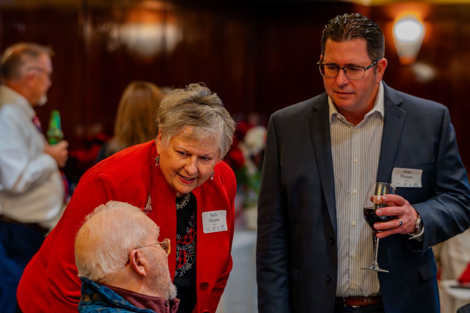 Three adults socializing at a formal event, woman in red jacket conversing with two men, one holding a glass of wine.