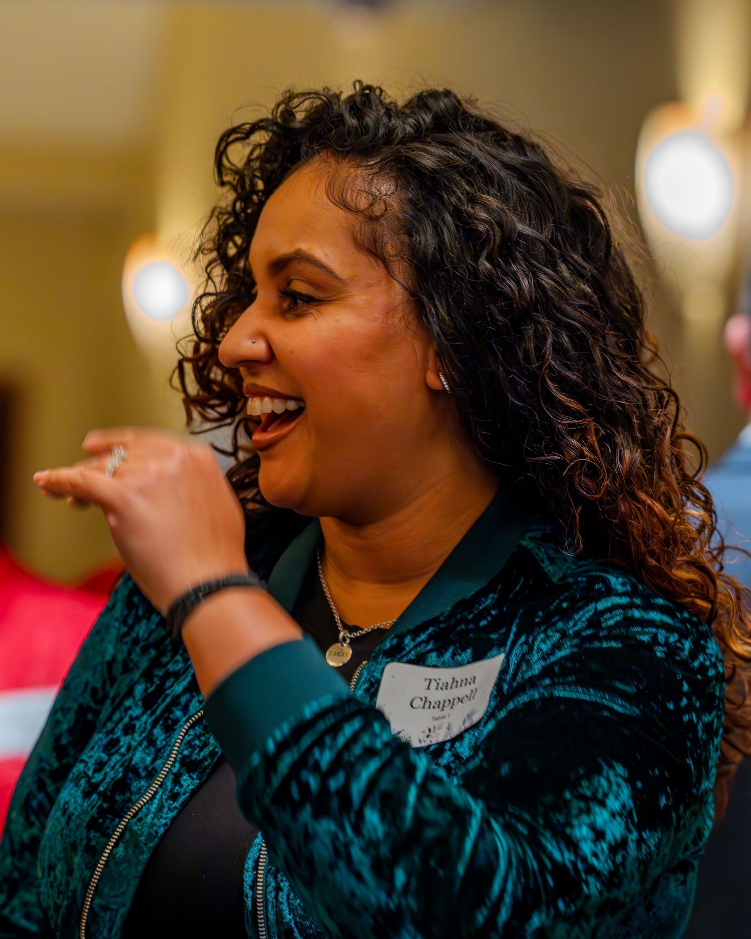 Smiling woman with curly hair wearing a teal patterned jacket and name tag at a networking event.