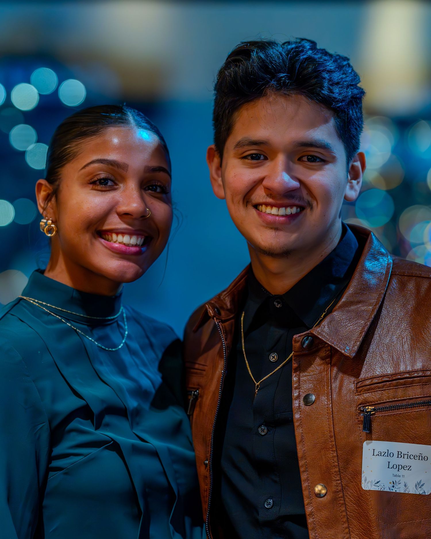 Two smiling young adults at a formal event, woman in teal dress and man in brown leather jacket wearing a name tag.