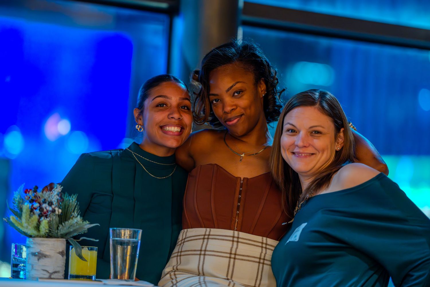Three women smiling and posing together at an indoor event with blue ambient lighting in the background.
