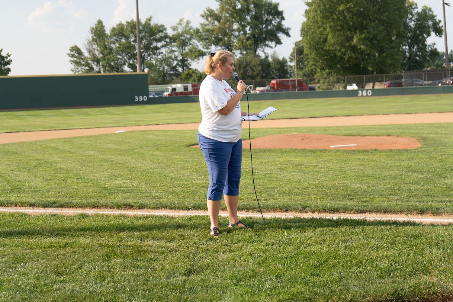 Woman singing into microphone on baseball field near pitcher's mound at outdoor stadium on sunny day.