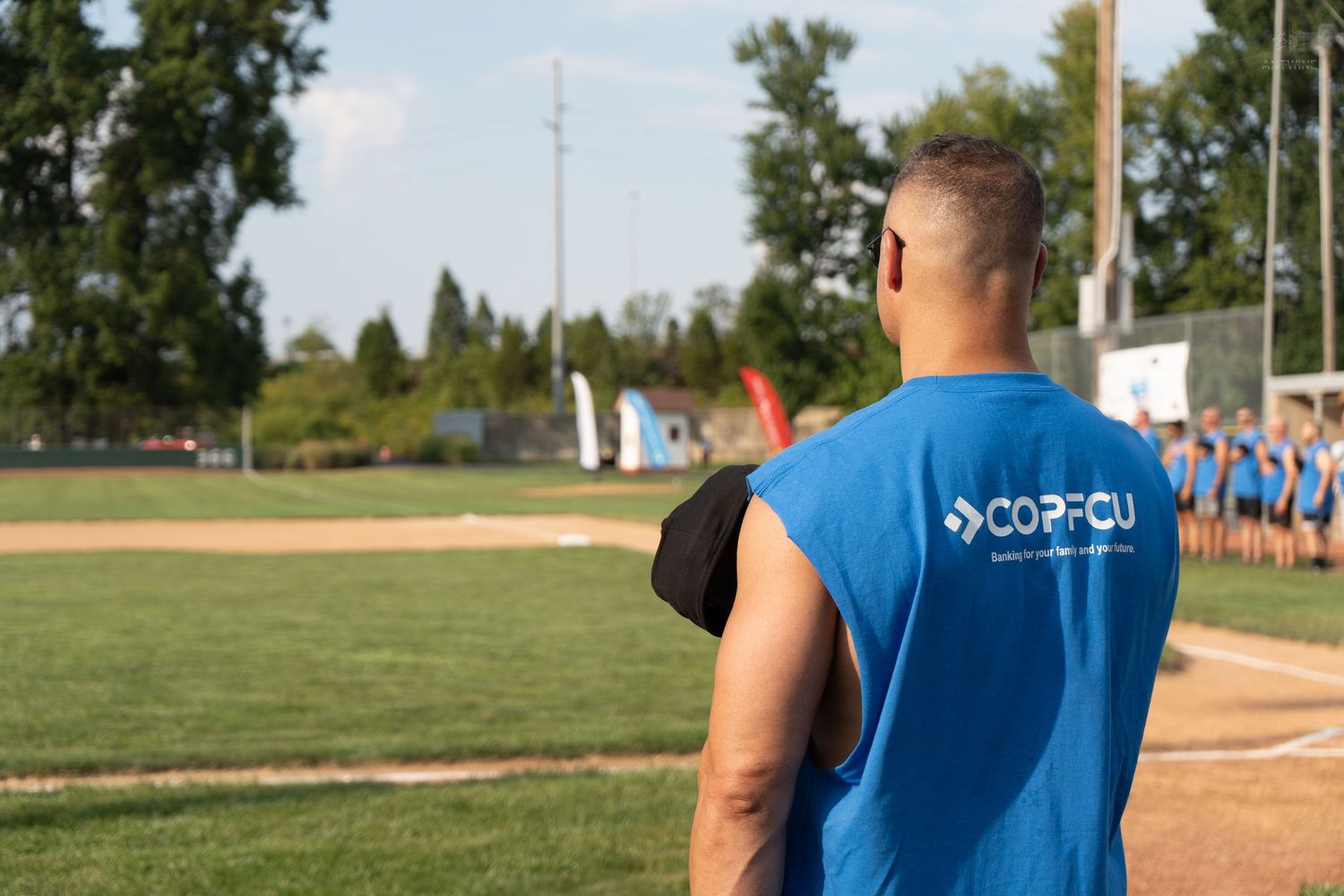 Man in blue COPFCU sleeveless shirt watching a baseball field event on a sunny day.