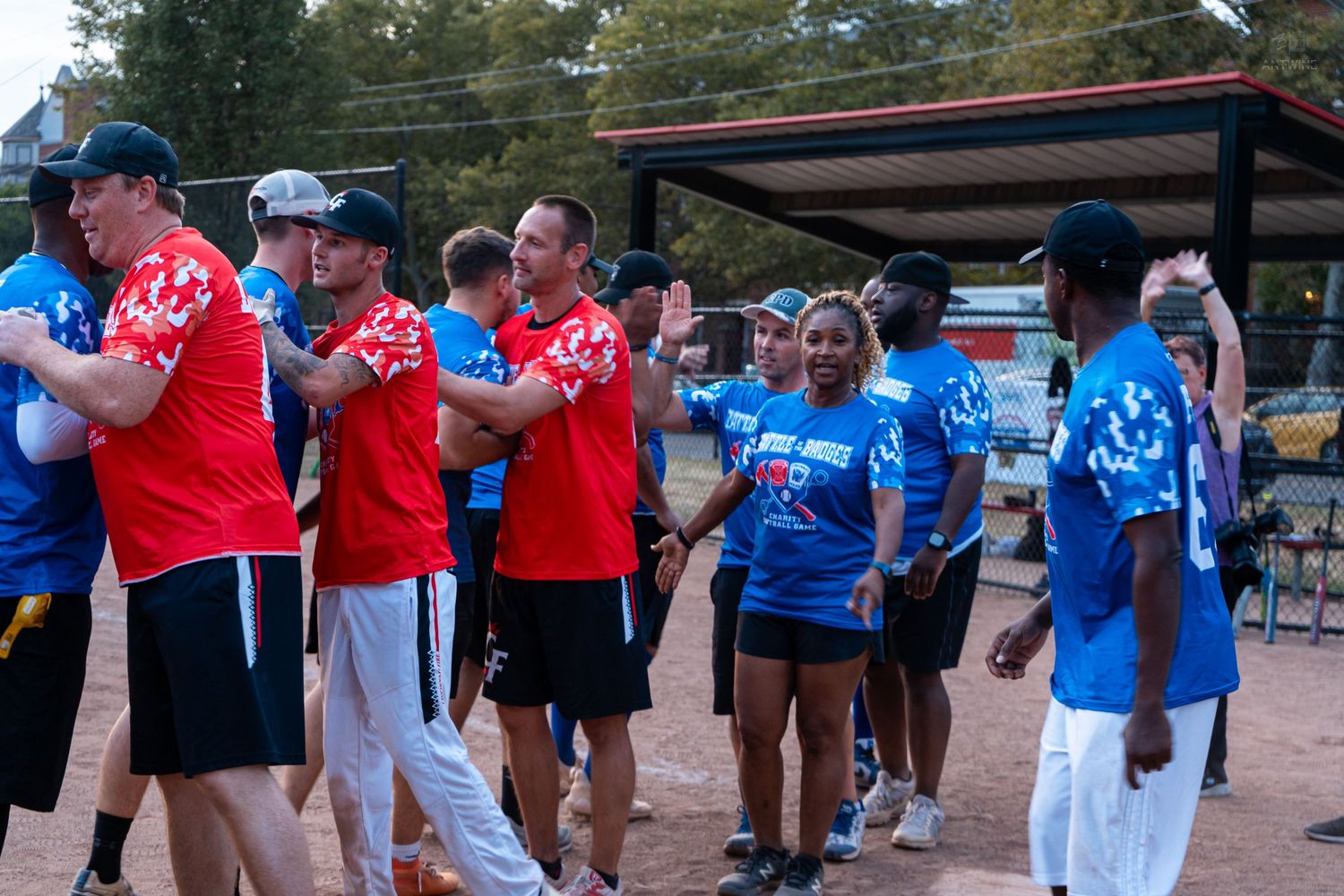 Teams in red and blue jerseys celebrate with high fives after a charity softball game.
