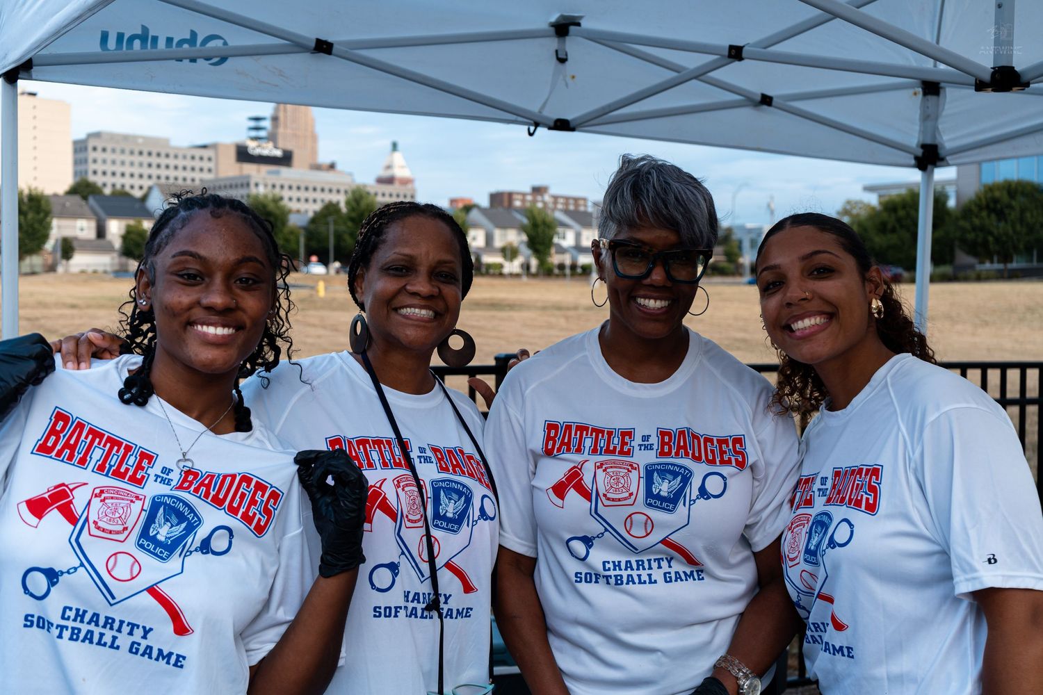 Four women smile together at a Battle of the Badges charity softball game under a tent.