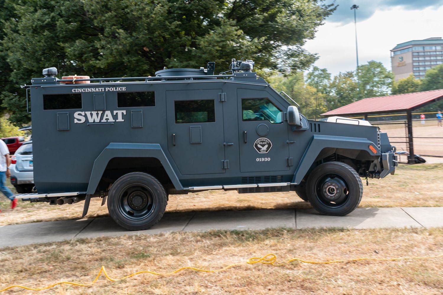 A black SWAT armored tactical police vehicle parked outdoors on grass near trees and a building.