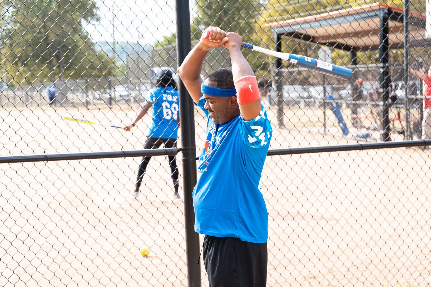 A softball player in a blue jersey stretches with a bat raised overhead behind a chain-link fence.