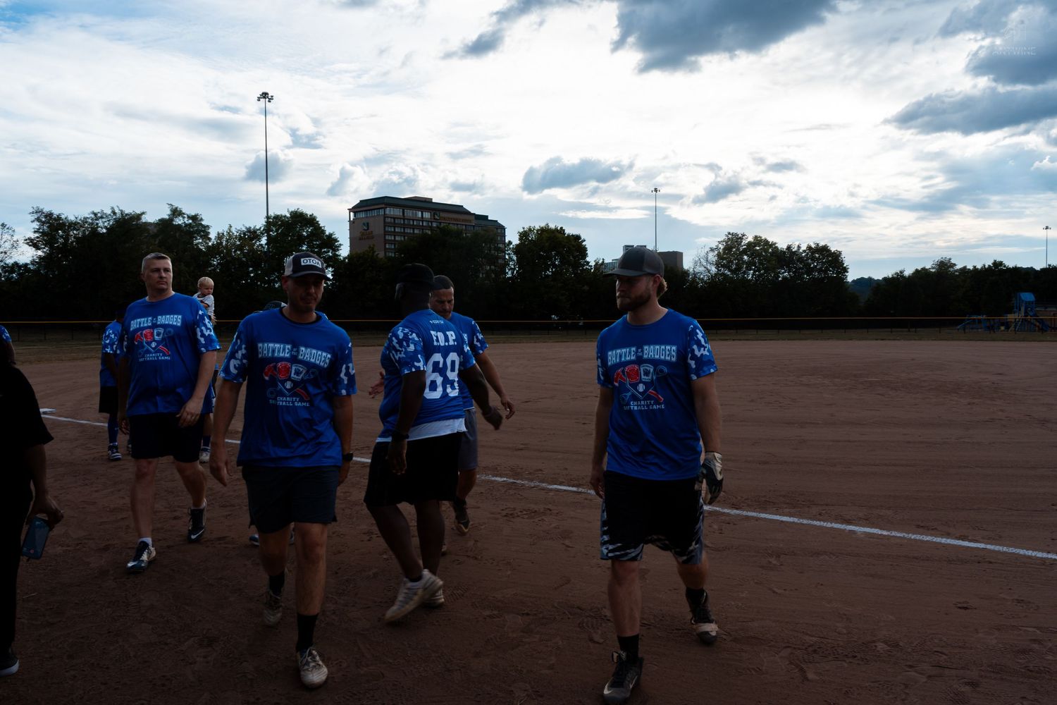 A group of men in matching blue softball jerseys walk across a dirt baseball field under a cloudy evening sky.