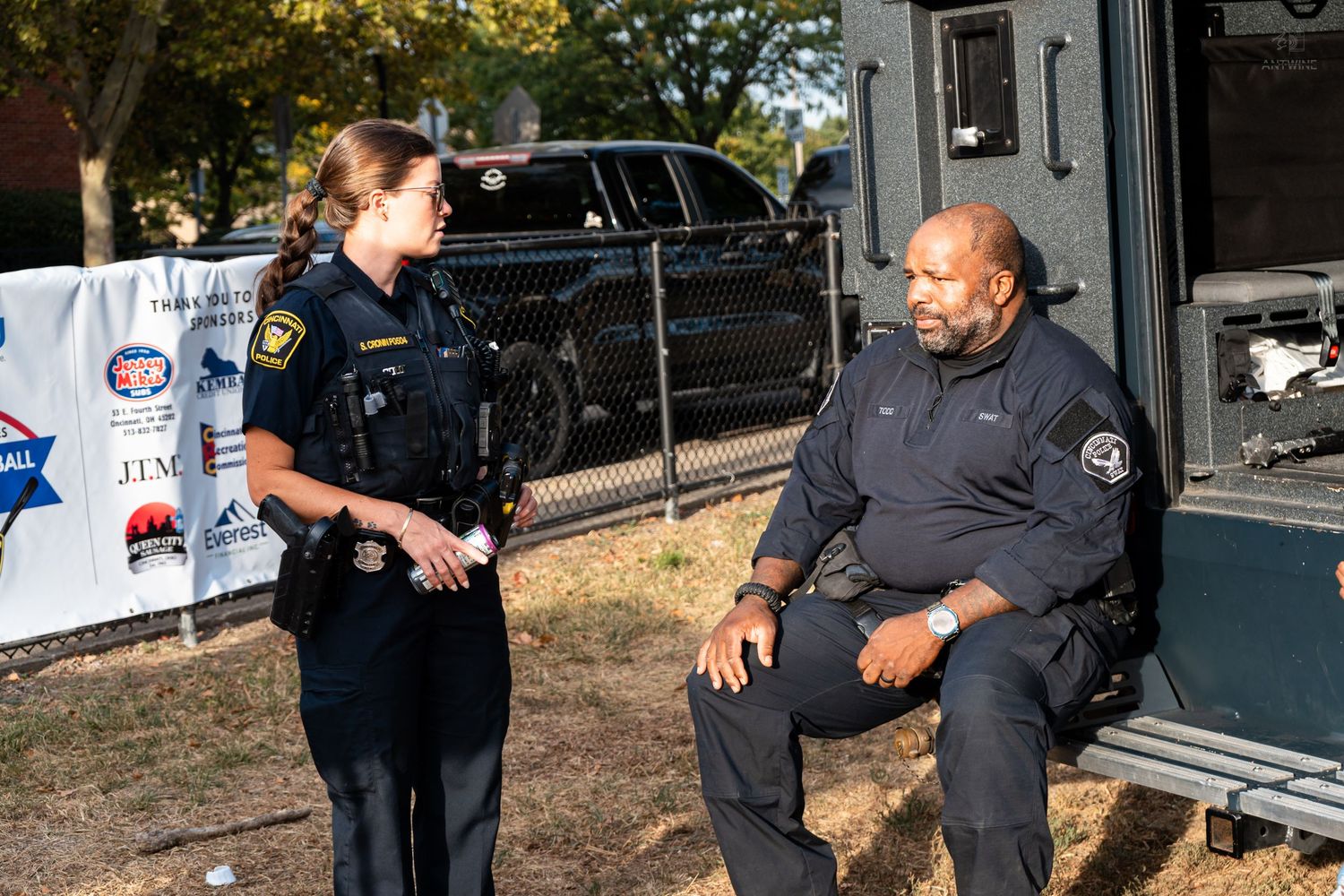 A female police officer in uniform converses with a security officer seated near a vehicle outdoors.