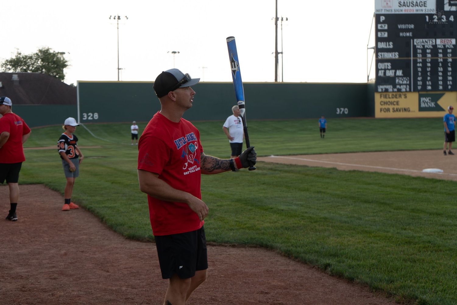 Man in red shirt holds a blue bat during an outdoor recreational softball game at a baseball stadium.