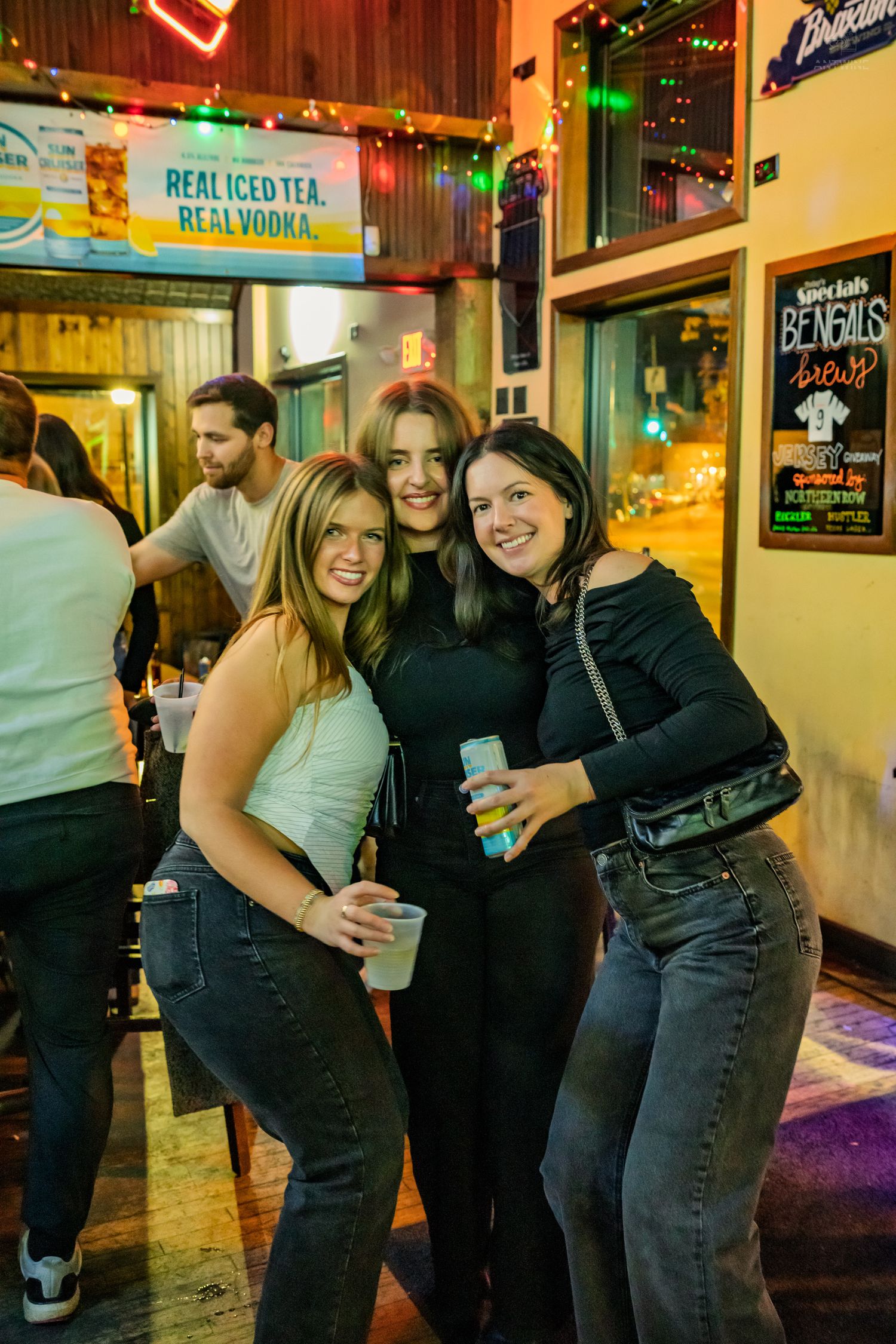 Three young women smiling and posing together at a lively bar, holding drinks, dressed in casual black and white outfits.