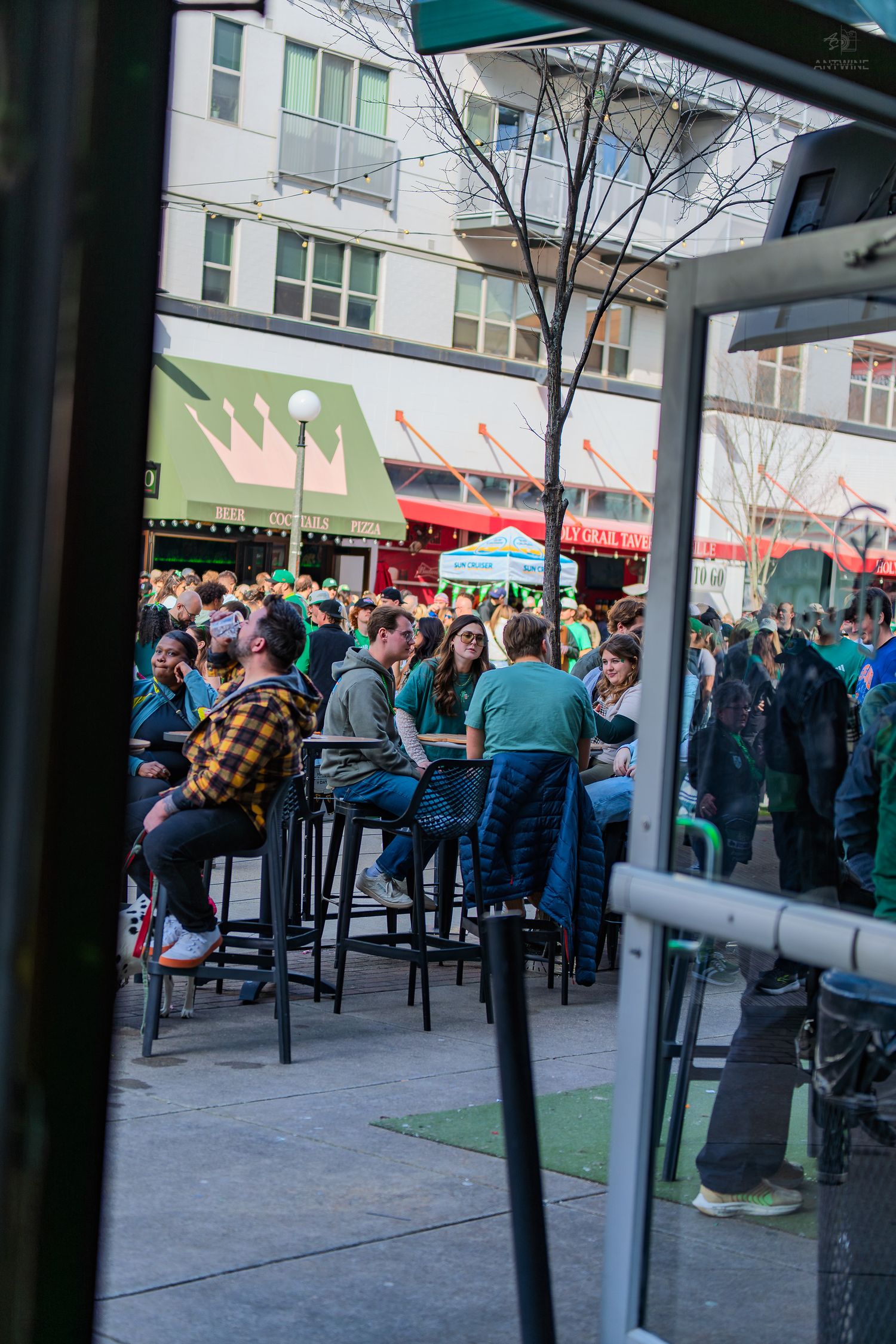 Crowded outdoor bar patio with people wearing green celebrating St. Patrick's Day in an urban setting.