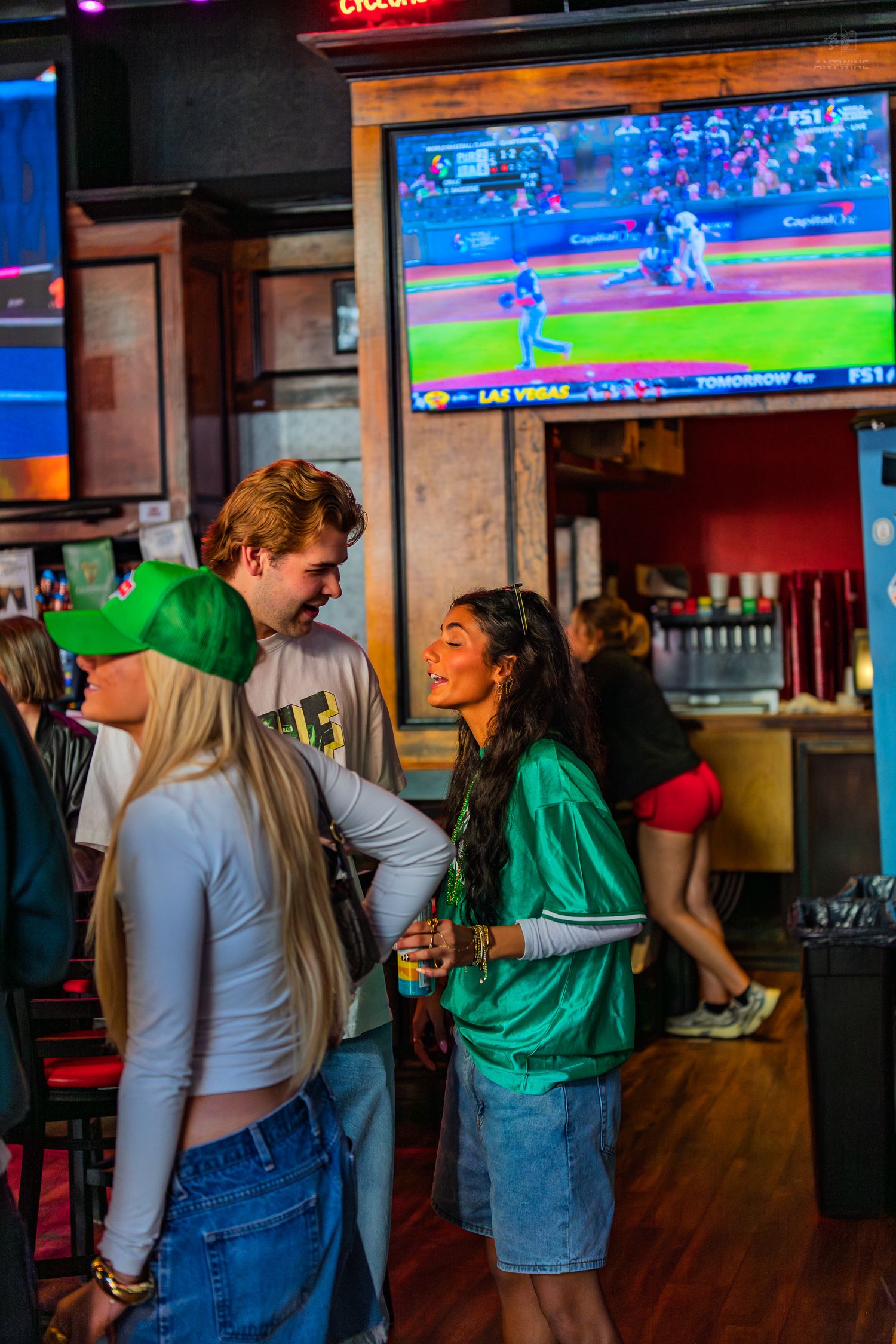 Friends in green outfits socializing at a sports bar with a baseball game on TV.