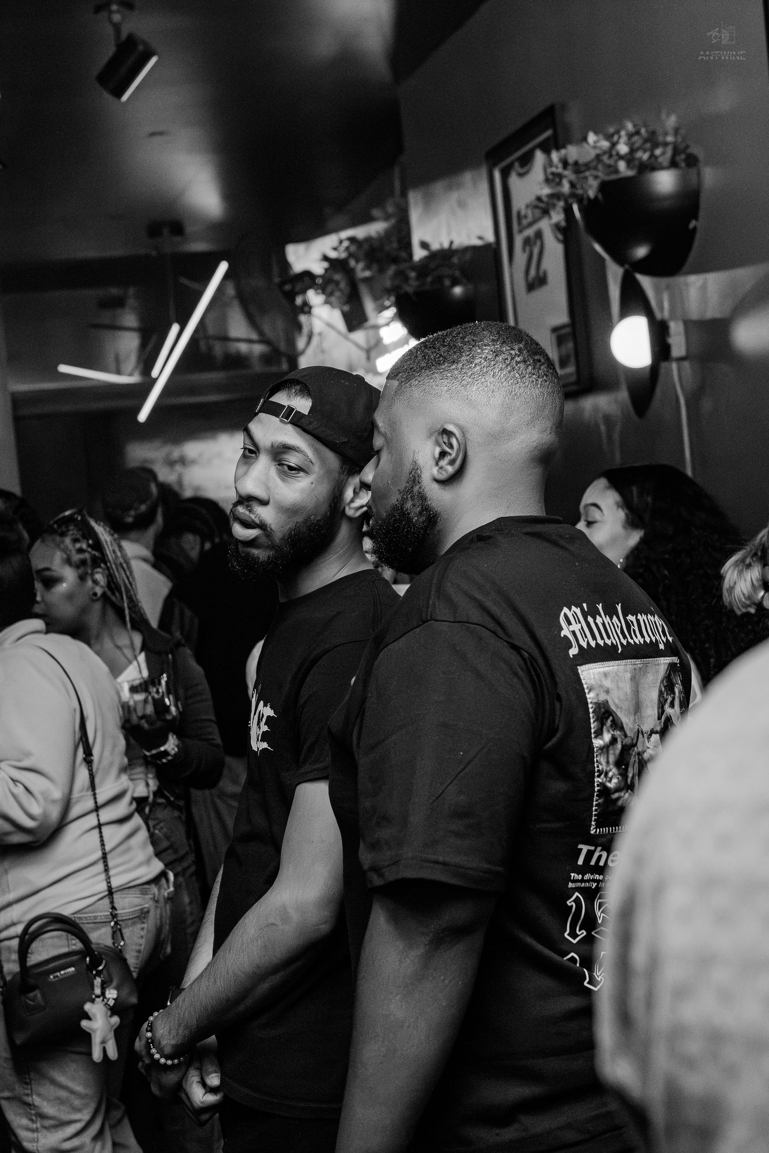 Two men in black shirts conversing at a crowded indoor social event, black and white.