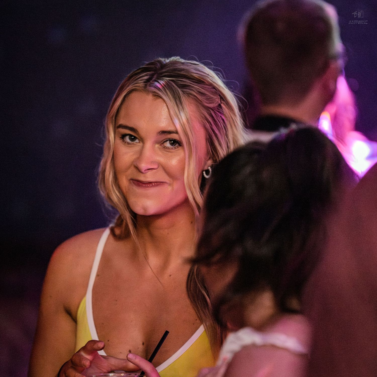 A smiling blonde woman in a yellow dress socializes at a dimly lit nightclub event with purple lighting.