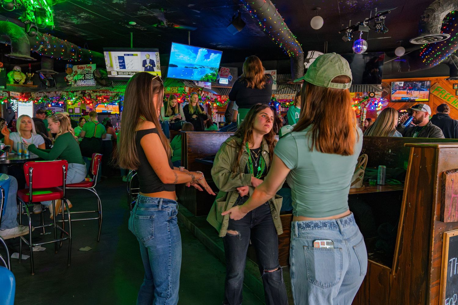 Women celebrating St. Patrick's Day at a festive bar with green decor, drinks, and TV screens in the background.