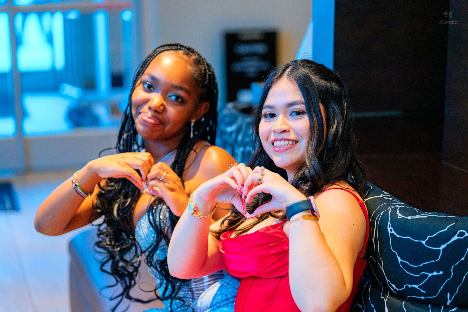 Two young women in formal gowns making heart shapes with their hands together, smiling at the camera.