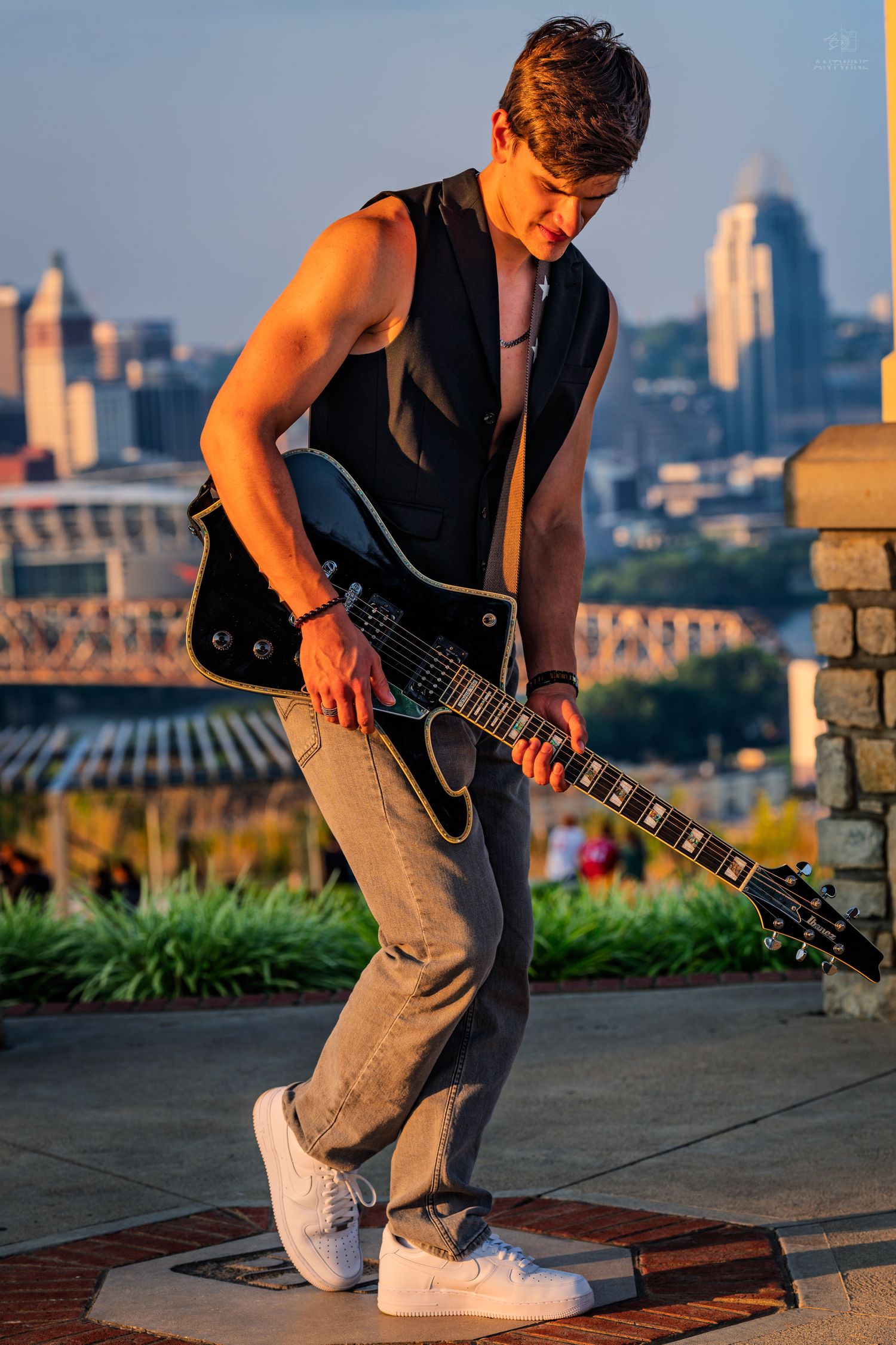 Young male guitarist playing black electric guitar outdoors with city skyline at sunset.
