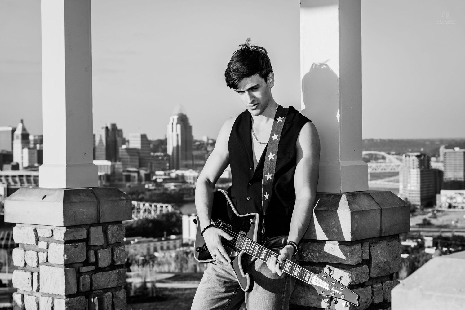 Young man playing electric guitar on rooftop with city skyline in black and white photo.