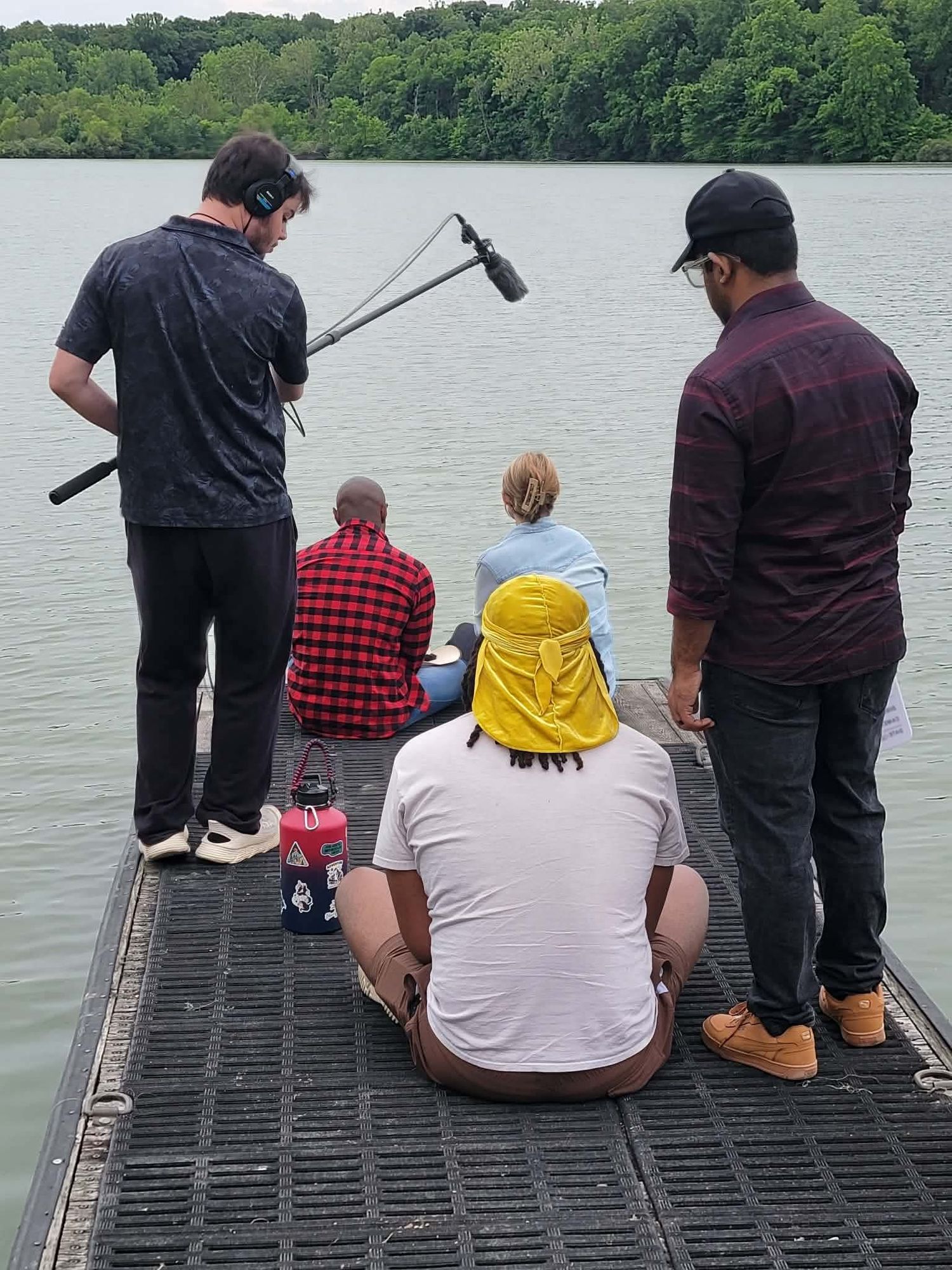 Film crew recording on a dock by a lake with two people sitting and one holding a boom microphone.