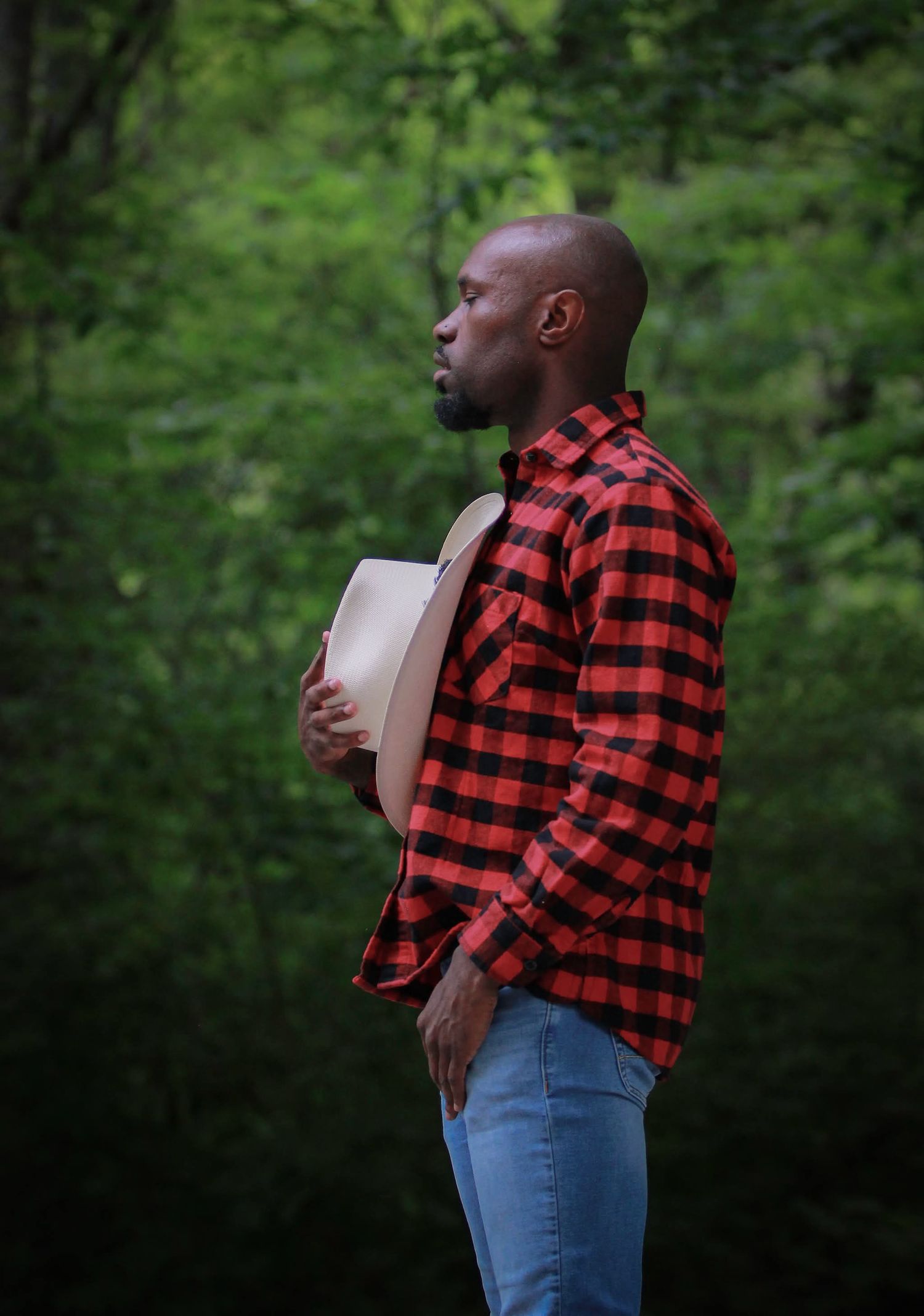 A bald Black man in a red plaid flannel shirt holds a hat while standing in a lush green forest.