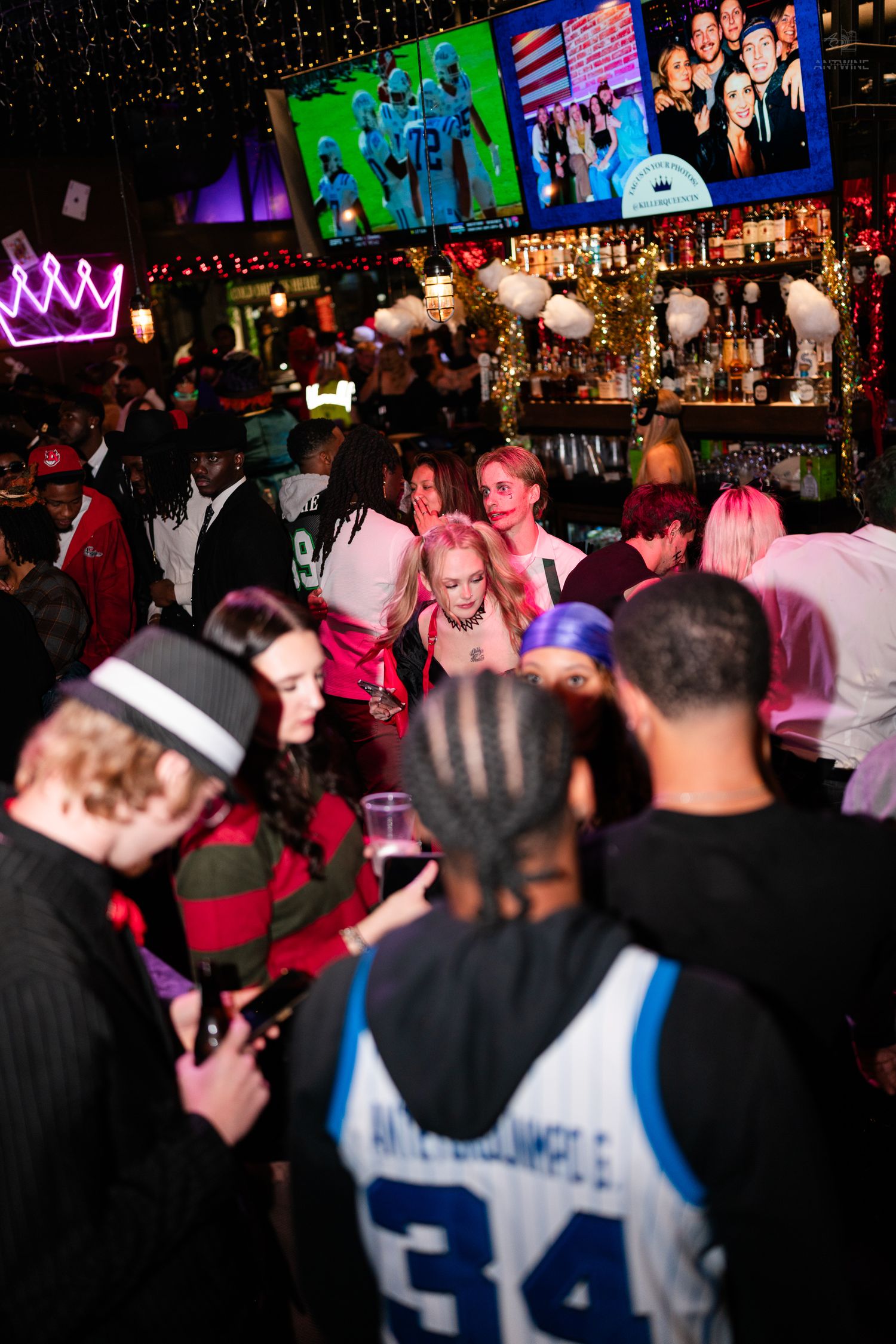 Crowded Halloween bar party with colorful costumes, TV screens, and festive decorations.