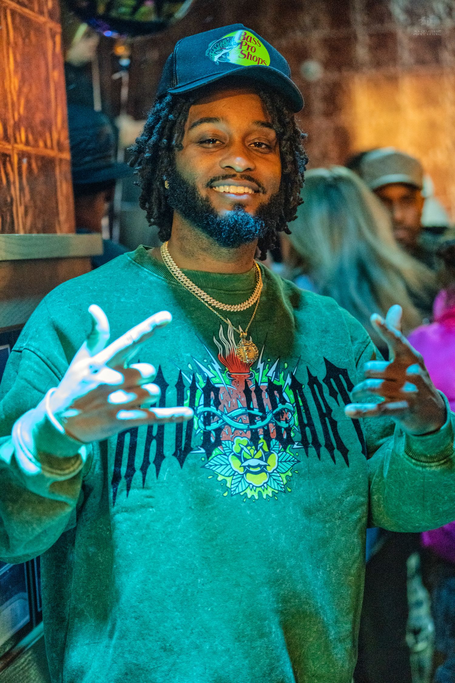 Smiling man in green sweatshirt and cap flashing peace signs at a lively indoor event.
