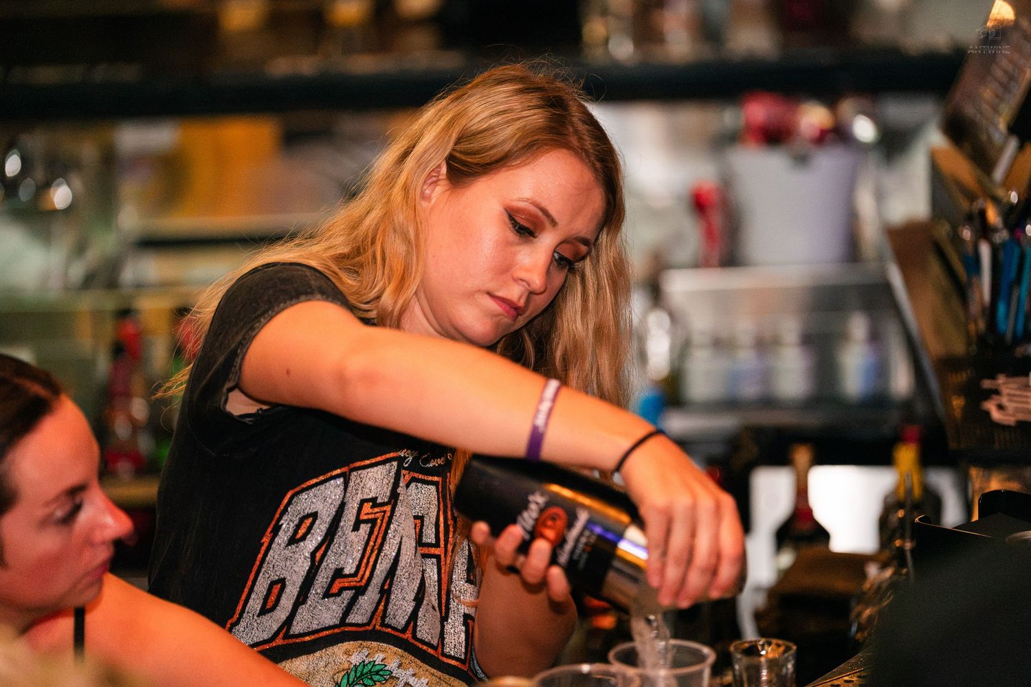 Female bartender pouring a drink behind a busy bar counter at a nightlife venue.