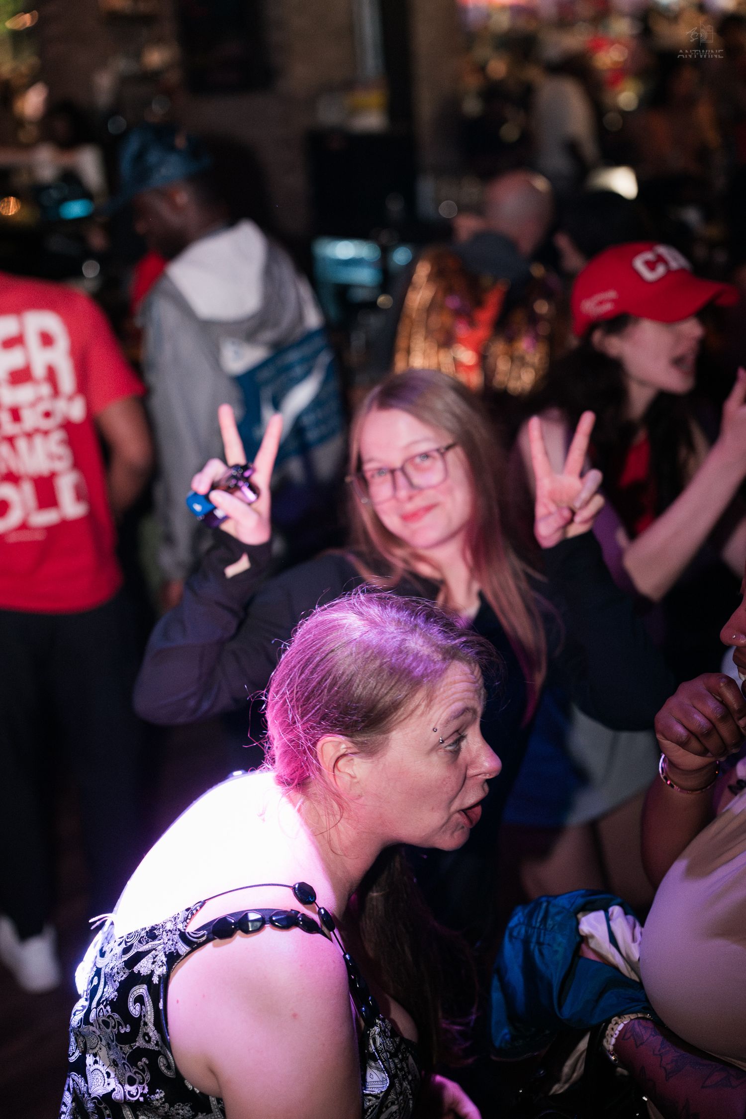 Woman with pink-highlighted hair dances as another flashes peace signs in a crowded nightclub.