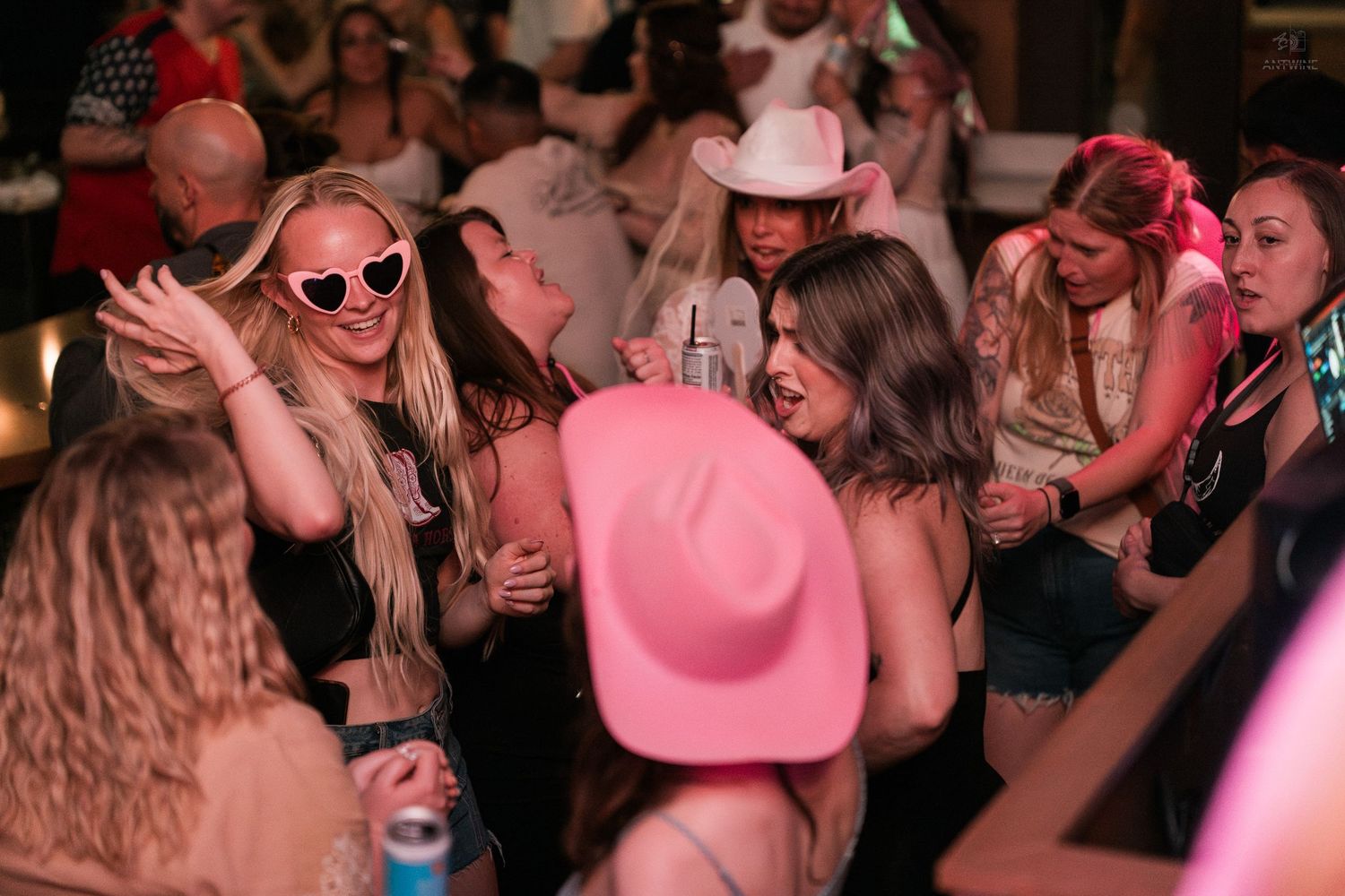 Group of women in pink cowboy hats and heart sunglasses dance together at a fun party.