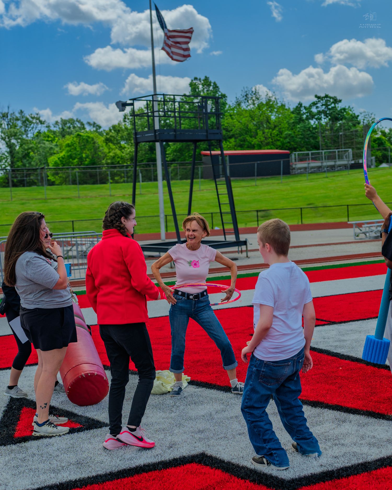 Students and teachers participate in an outdoor activity on a red track field with an American flag in the background.