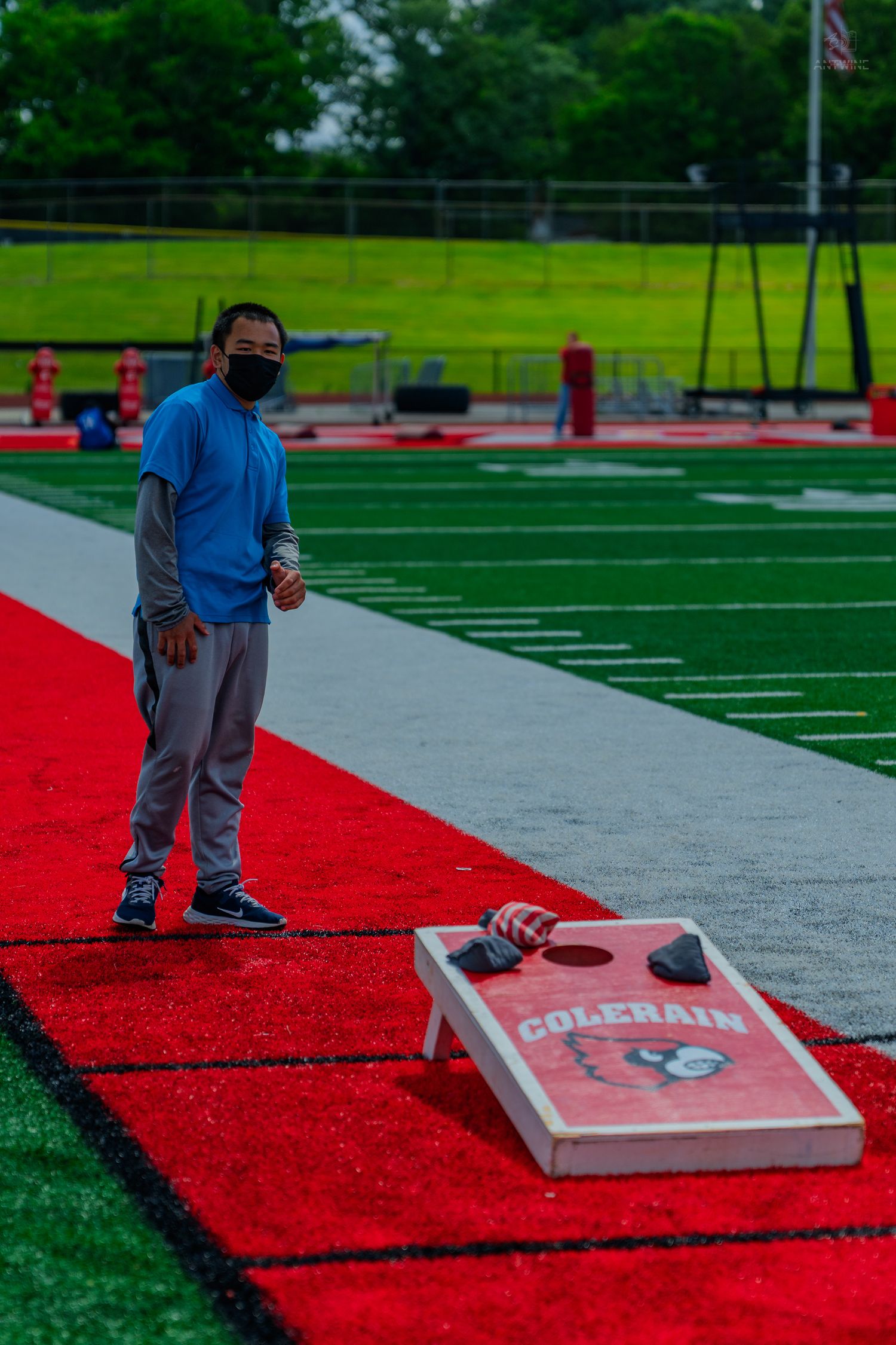 Man in blue shirt and mask playing cornhole on red turf at Colerain football field.