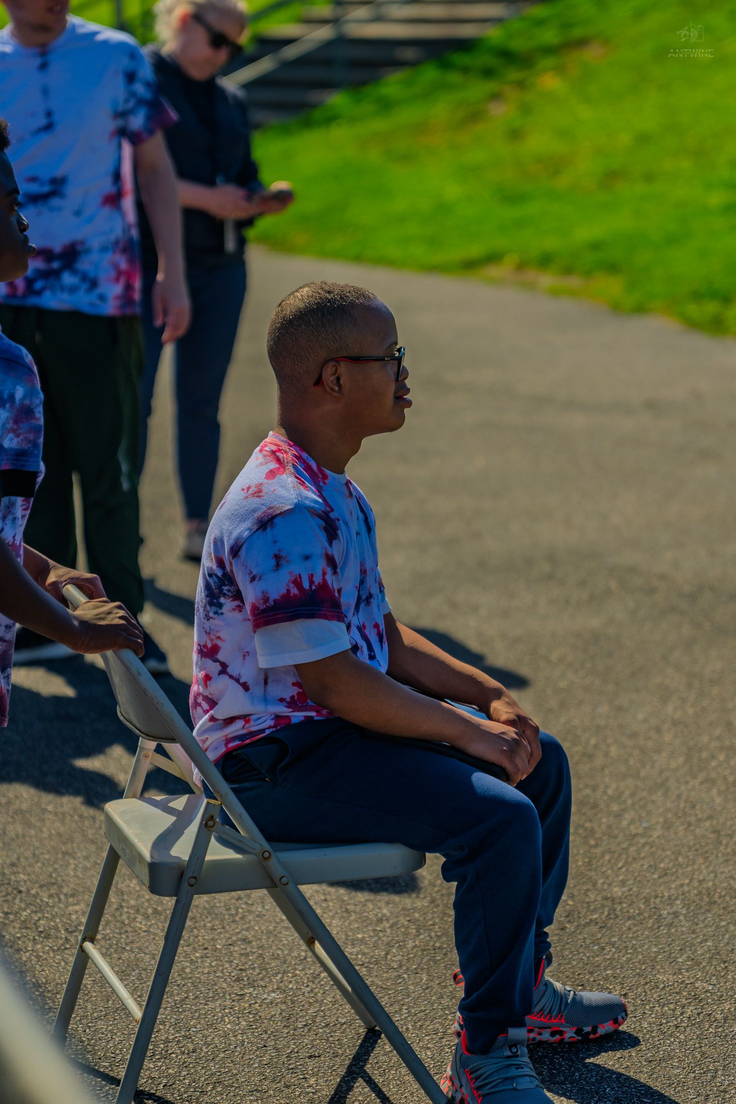 Young man in tie-dye shirt sitting on folding chair outdoors, wearing glasses, with people standing behind him.