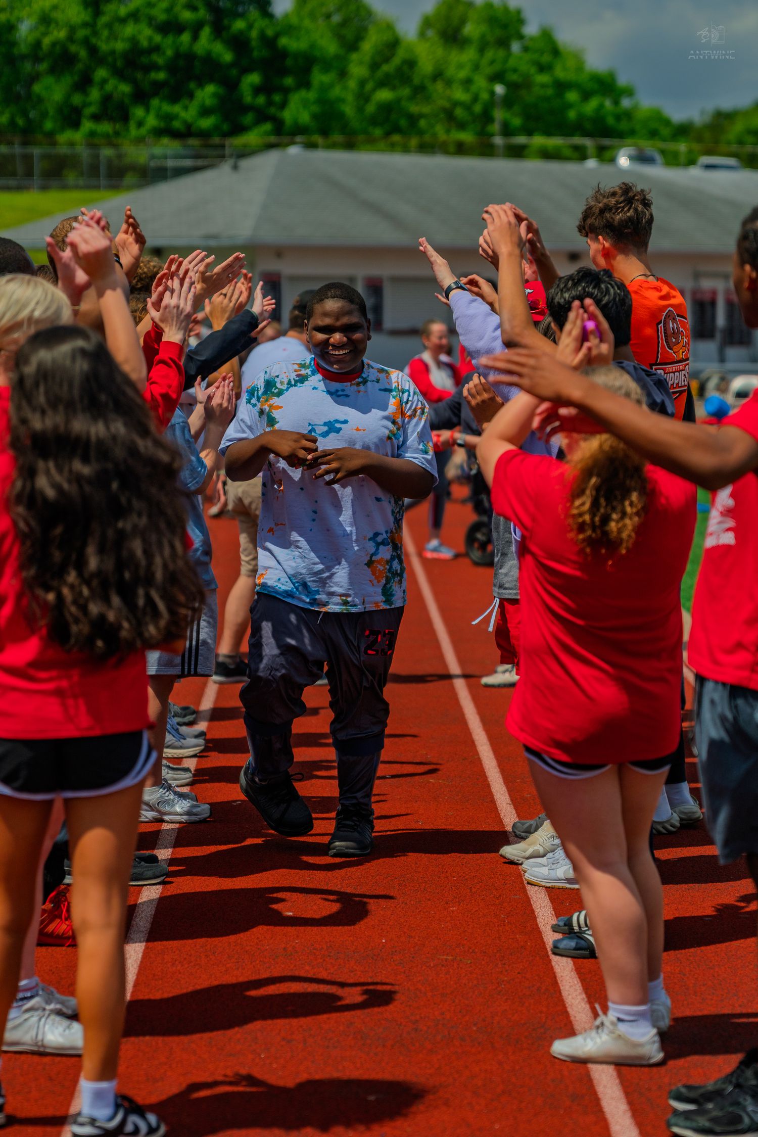 Students cheering on a peer running through a celebratory tunnel on a red track at an outdoor school sports event.
