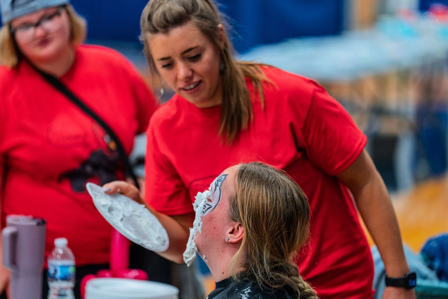 A woman in a red shirt applies face paint to a child at a festive outdoor event.