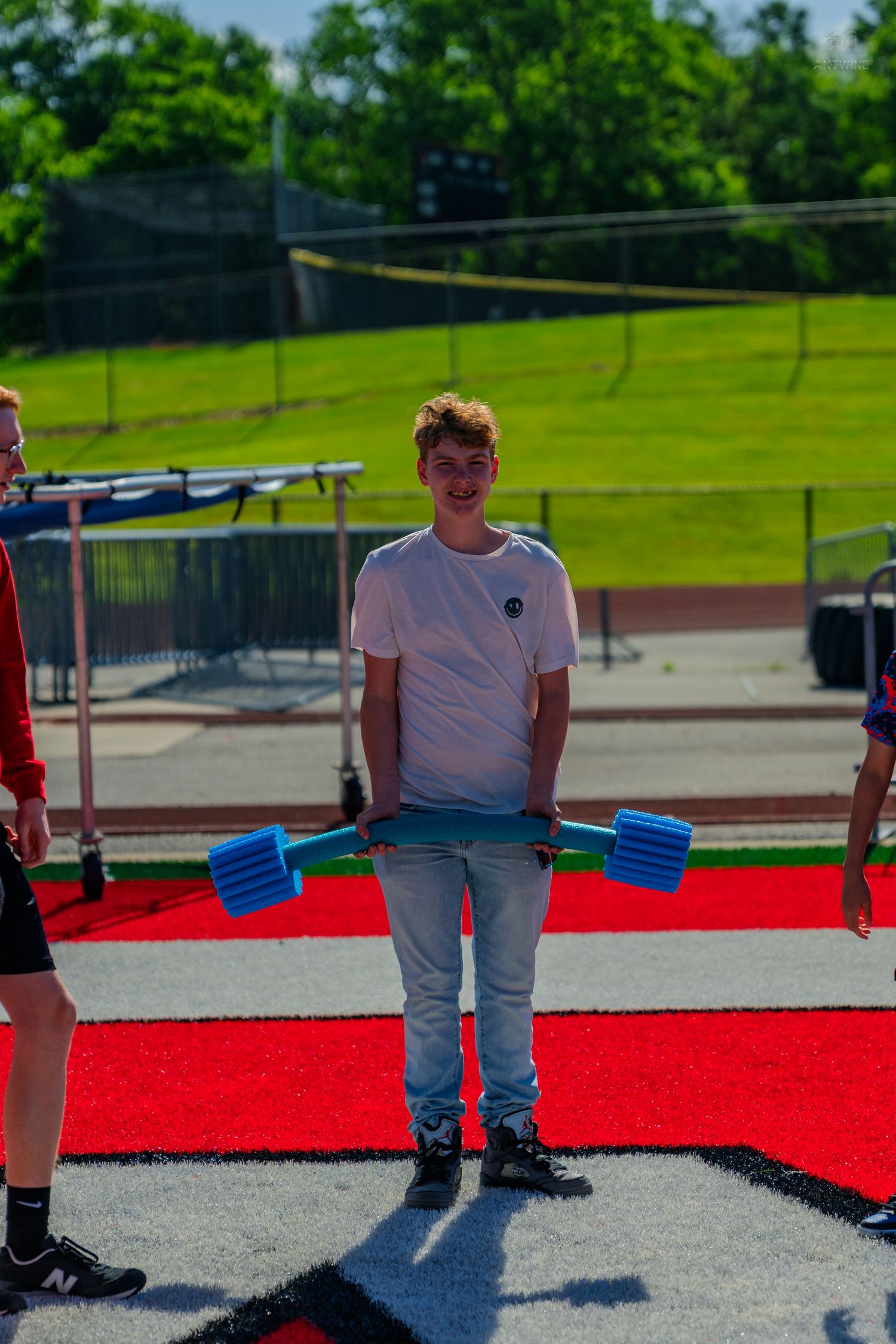 A smiling teen boy holds blue foam dumbbells on a red track surface outdoors on a sunny day.