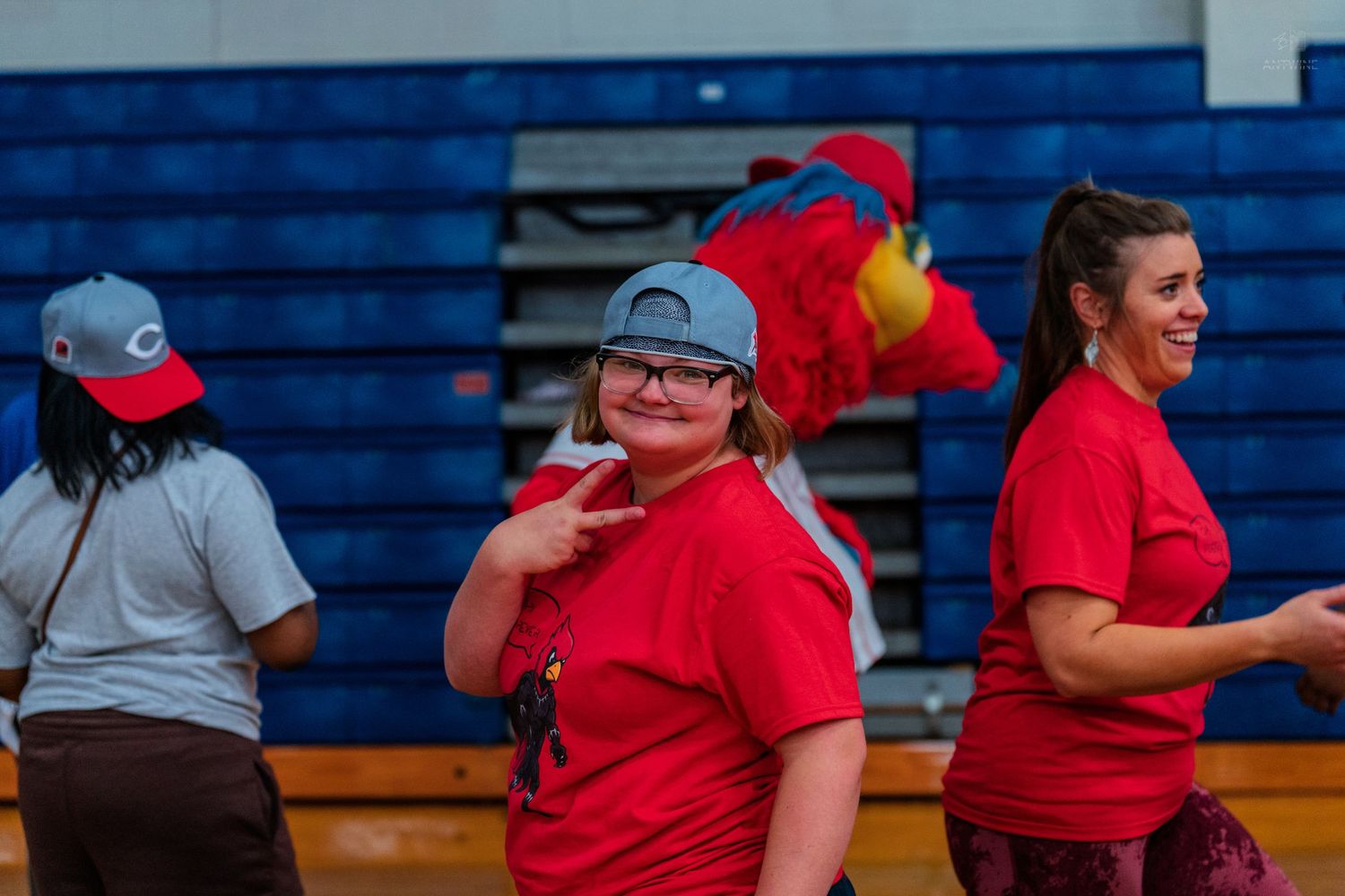 A young girl in a red t-shirt poses with a peace sign in a gymnasium with a mascot visible behind her.