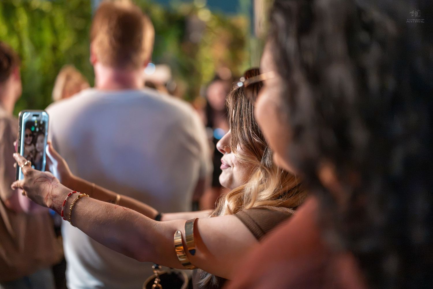 Woman taking a selfie with smartphone at outdoor festival, wearing bracelets, crowd visible in background.