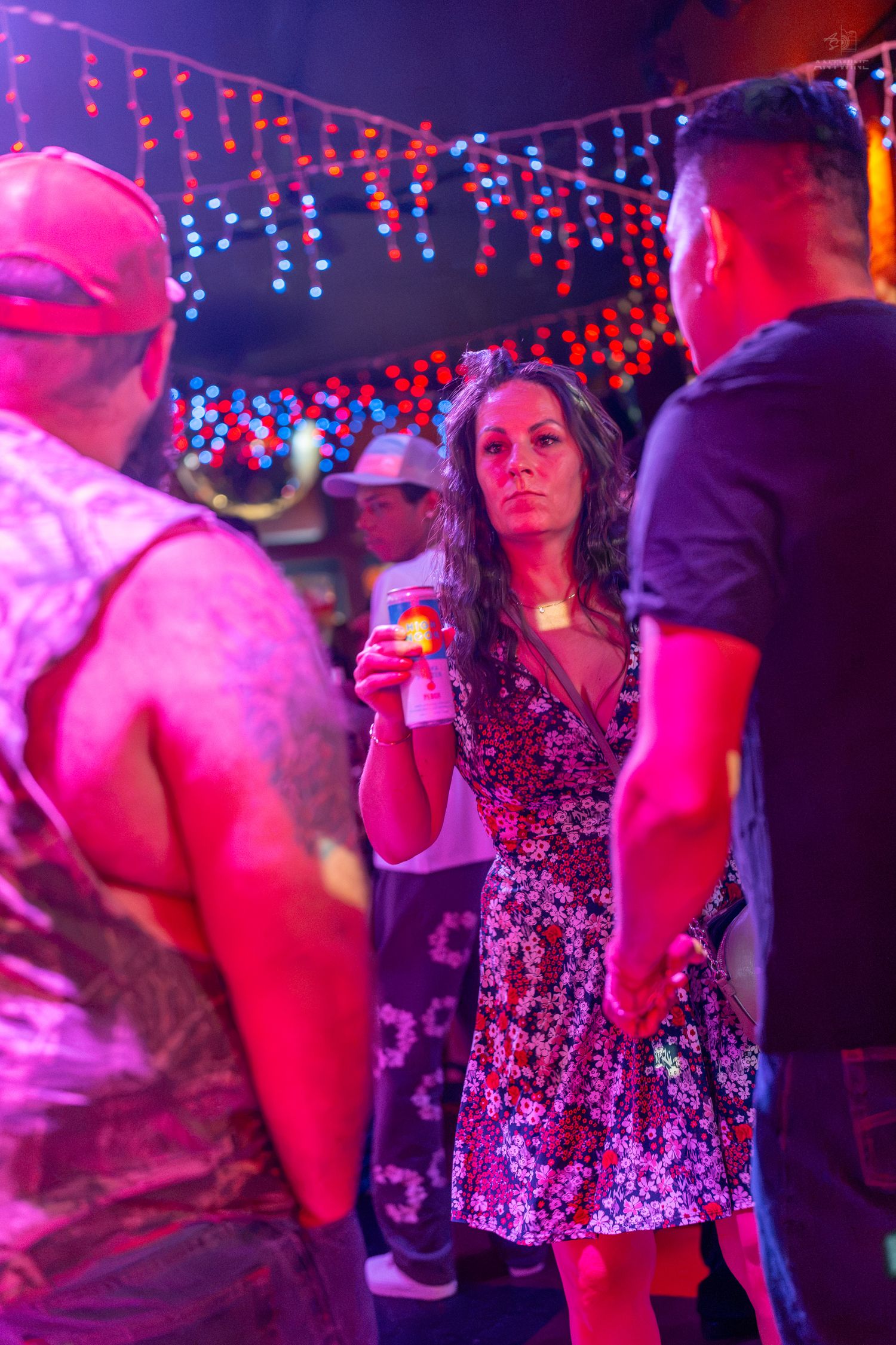 Woman in floral dress holding cocktail at vibrant neon-lit nightclub party with colorful string lights.