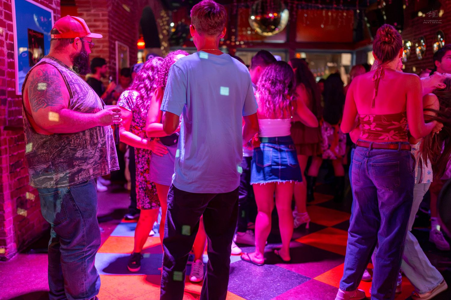 People dancing in a vibrant nightclub with colorful purple and pink lighting on a checkered dance floor.
