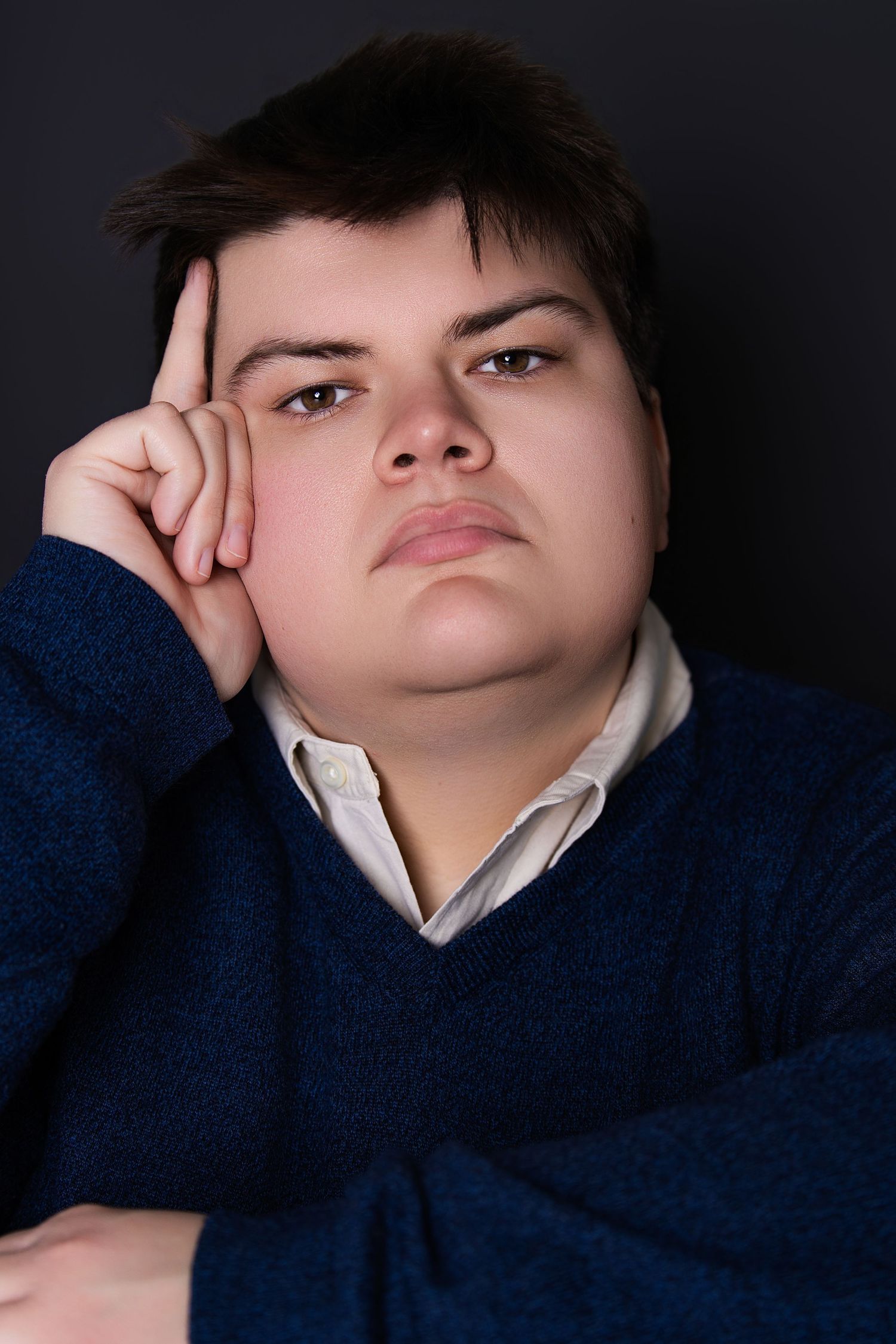 Color portrait of a young person in blue sweater resting head on hand against dark background.
