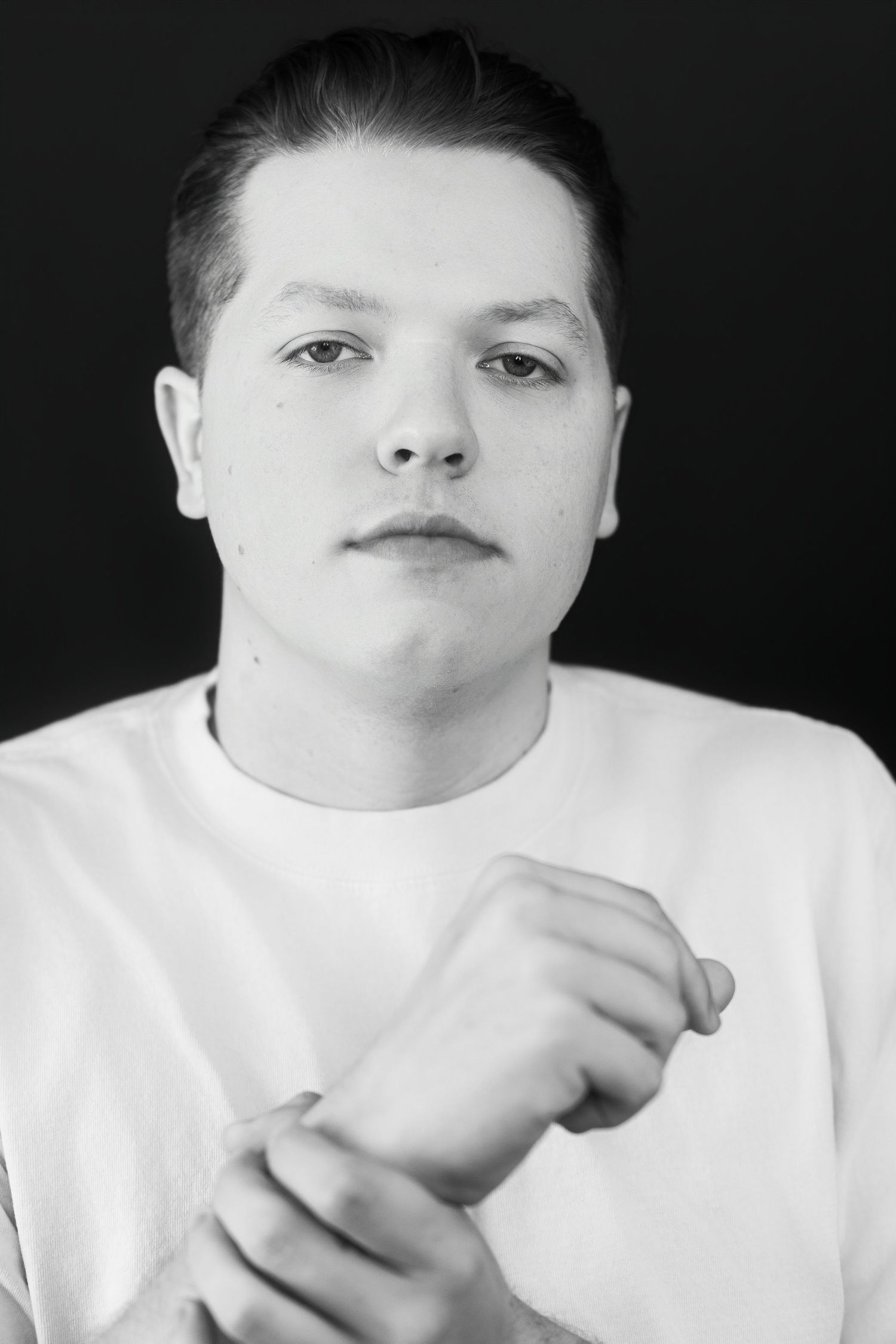 Black and white portrait of young man in white t-shirt holding wrist, direct gaze.