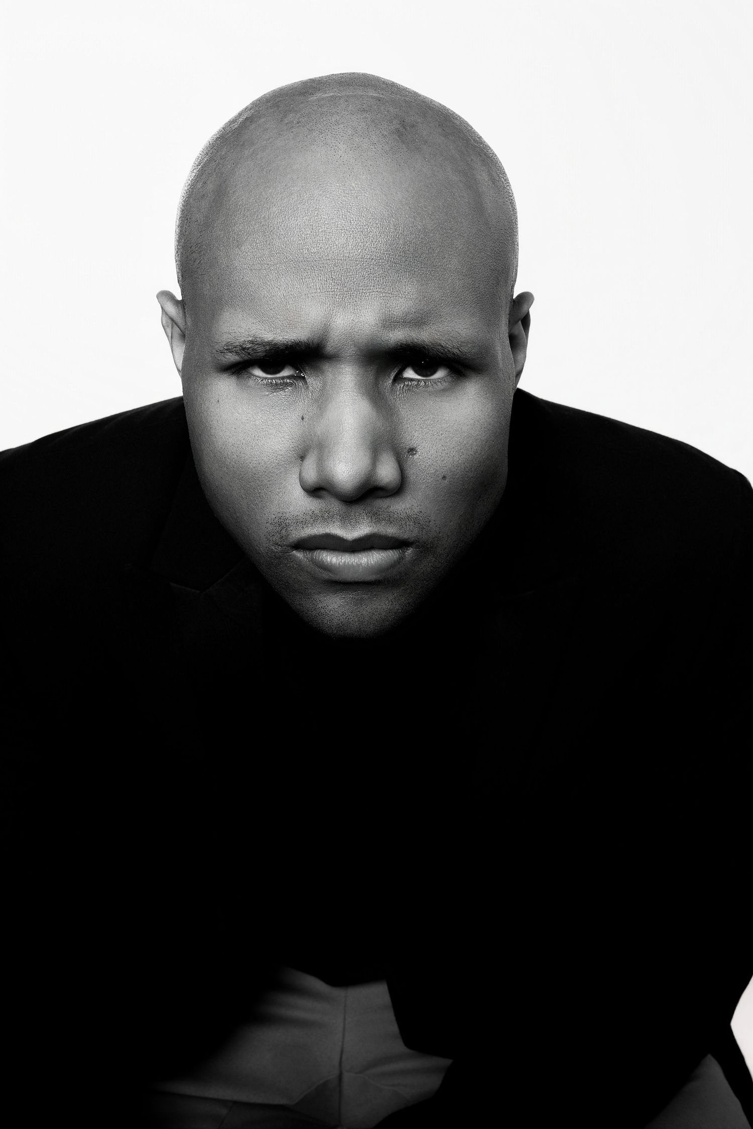 Bald Black man in black shirt with serious expression in black and white portrait photo.