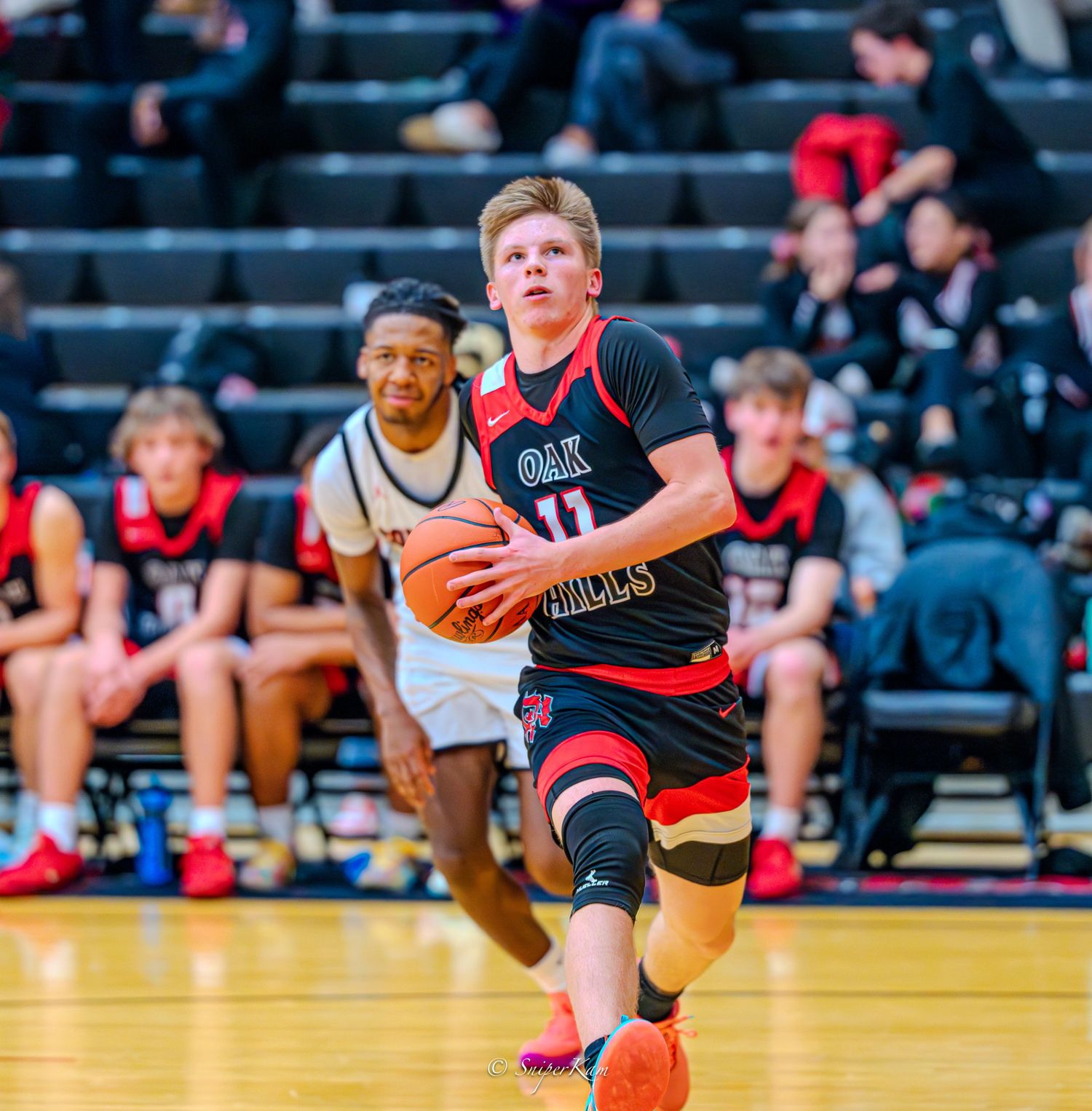 High school basketball player in Oak Hills jersey dribbling down court during competitive game.