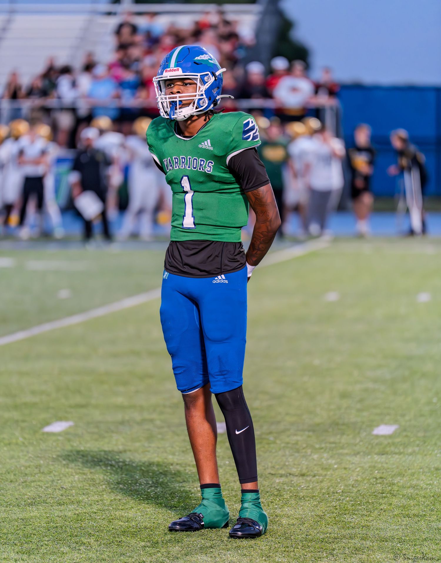Warriors football player #1 in green and blue uniform stands on the field during a daytime game.