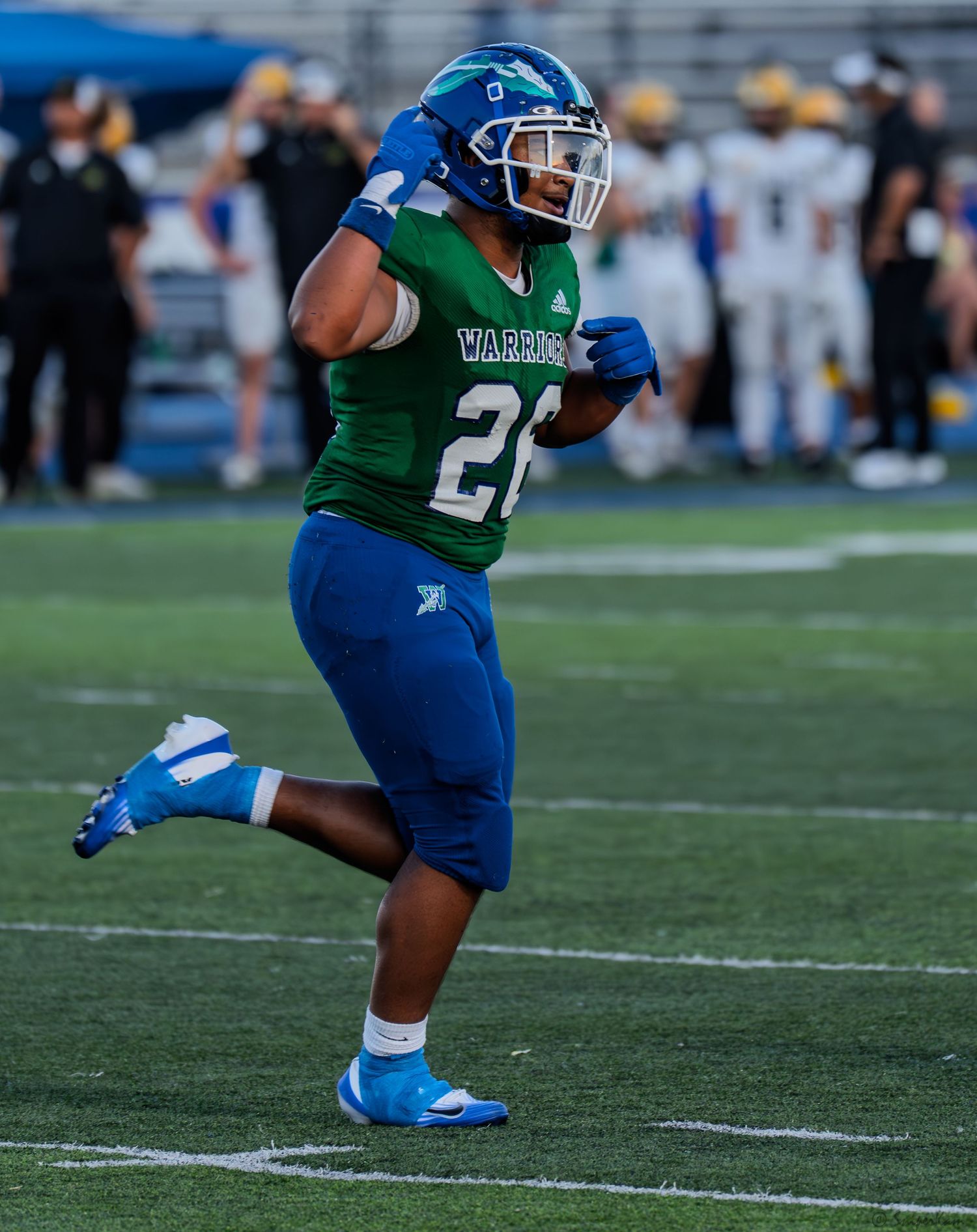 Warriors football player #26 in green and blue uniform celebrates on field during high school game.