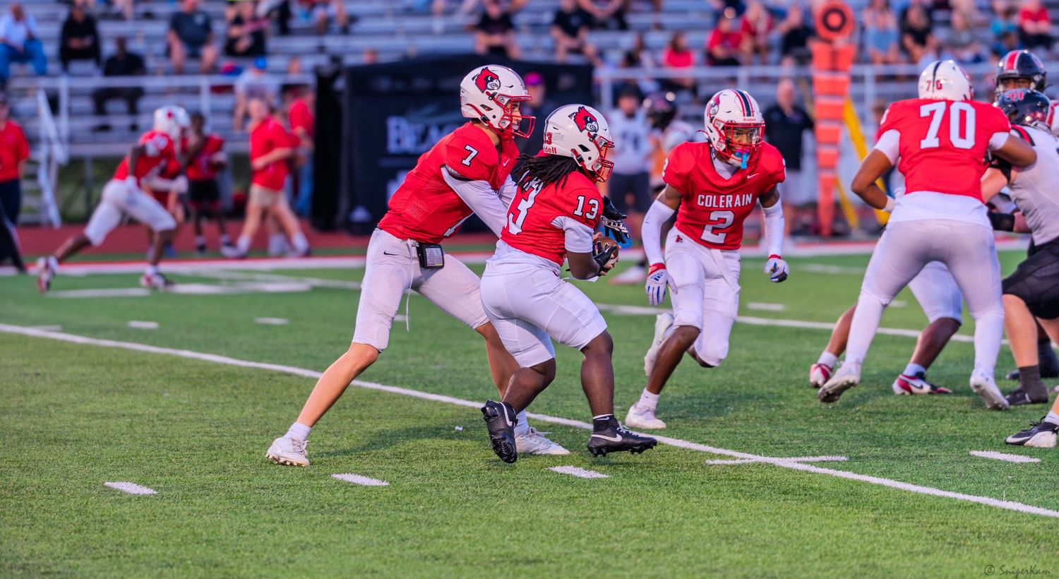 Red-uniformed quarterback #7 hands off the football to a teammate during an offensive play in a high school football game.