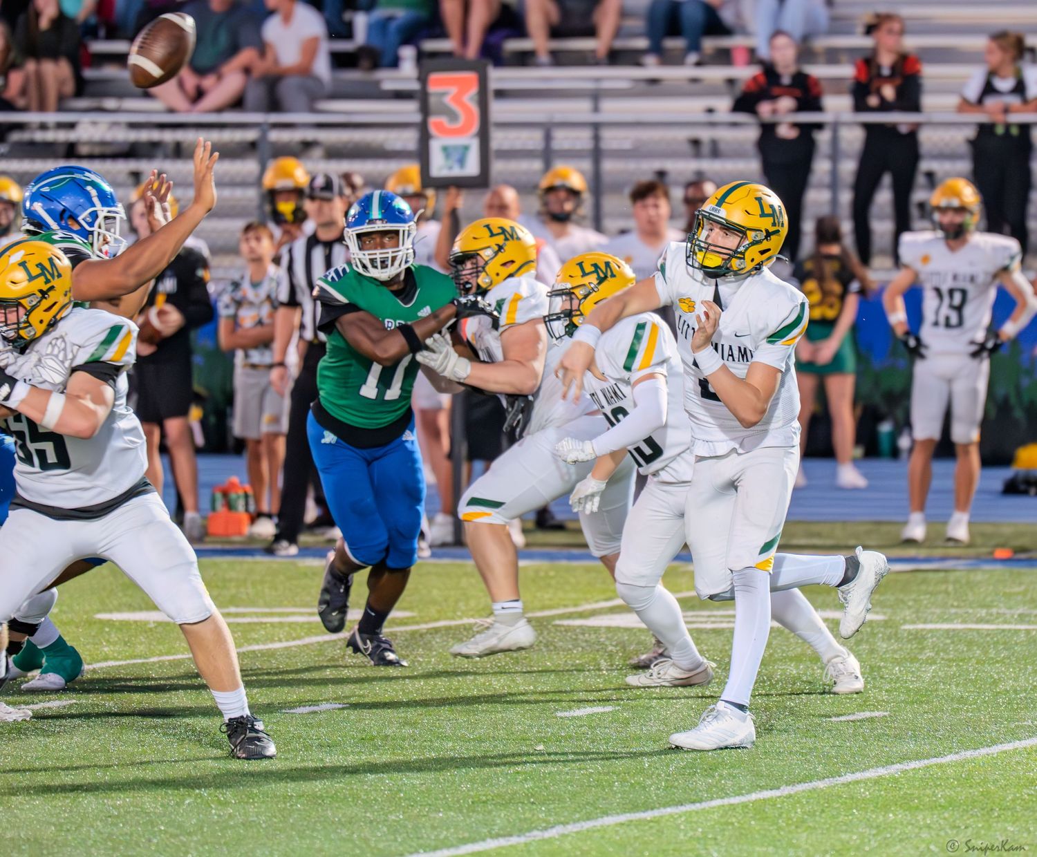 A quarterback in white and gold uniform releases a pass while under pressure during a high school football game.
