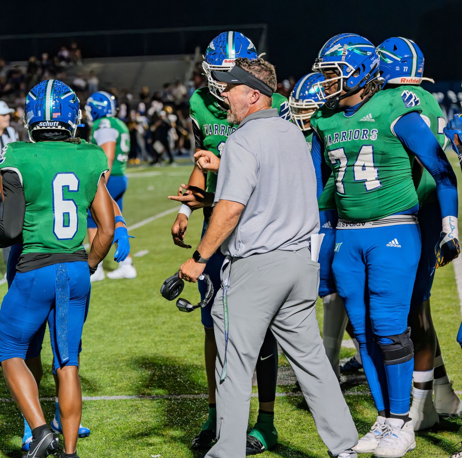 Warriors football coach in gray talks to player number 74 on the sideline during a game.
