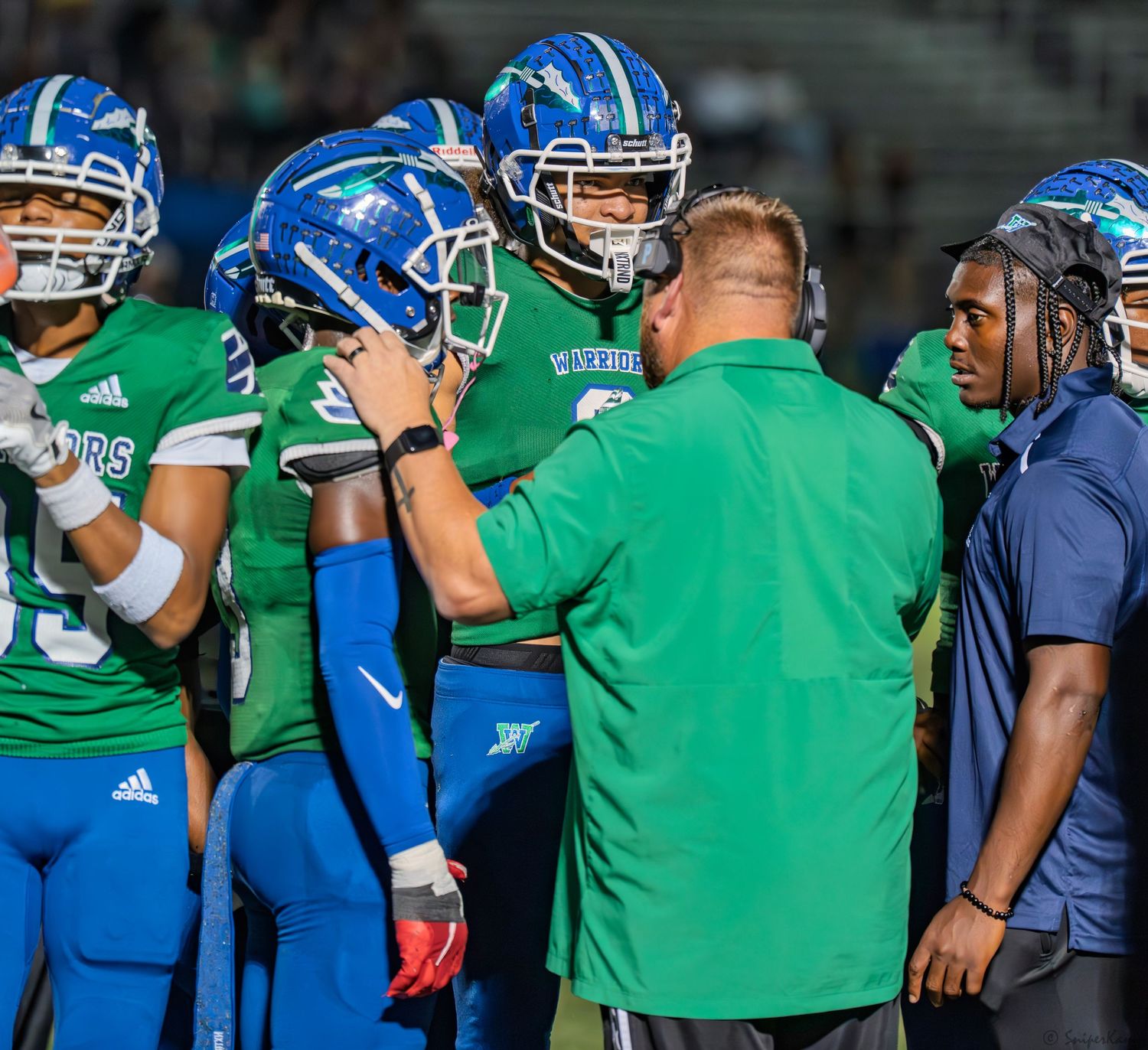 High school football coach in green jacket huddles with players in blue and green uniforms on the sideline.